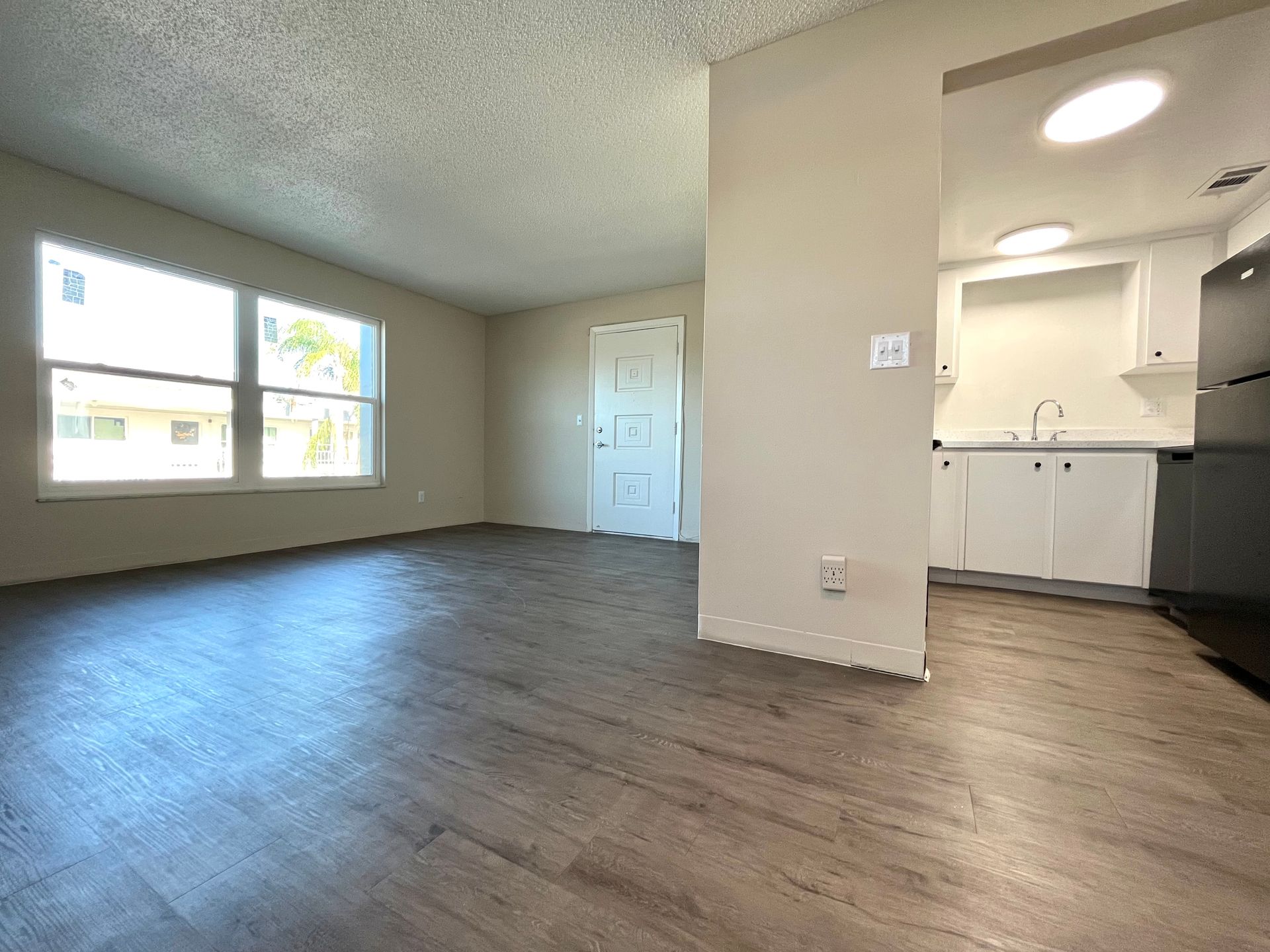 Empty living space with gray wood-look flooring, white walls, large windows, and a small kitchen area.