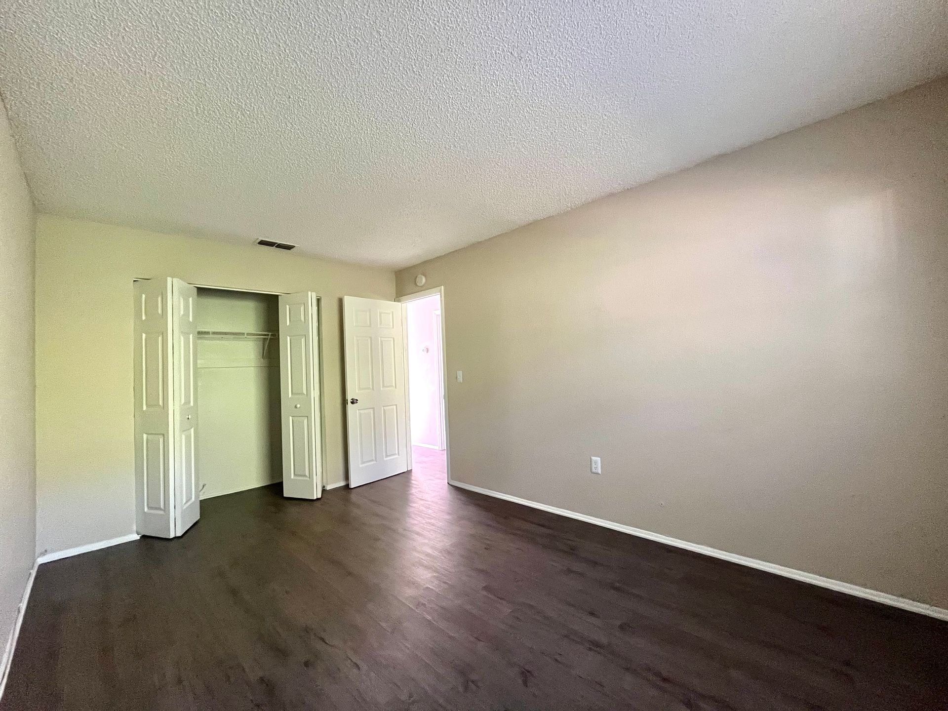 Empty bedroom with dark wood floors, closet, and open door. Light tan walls and textured ceiling.