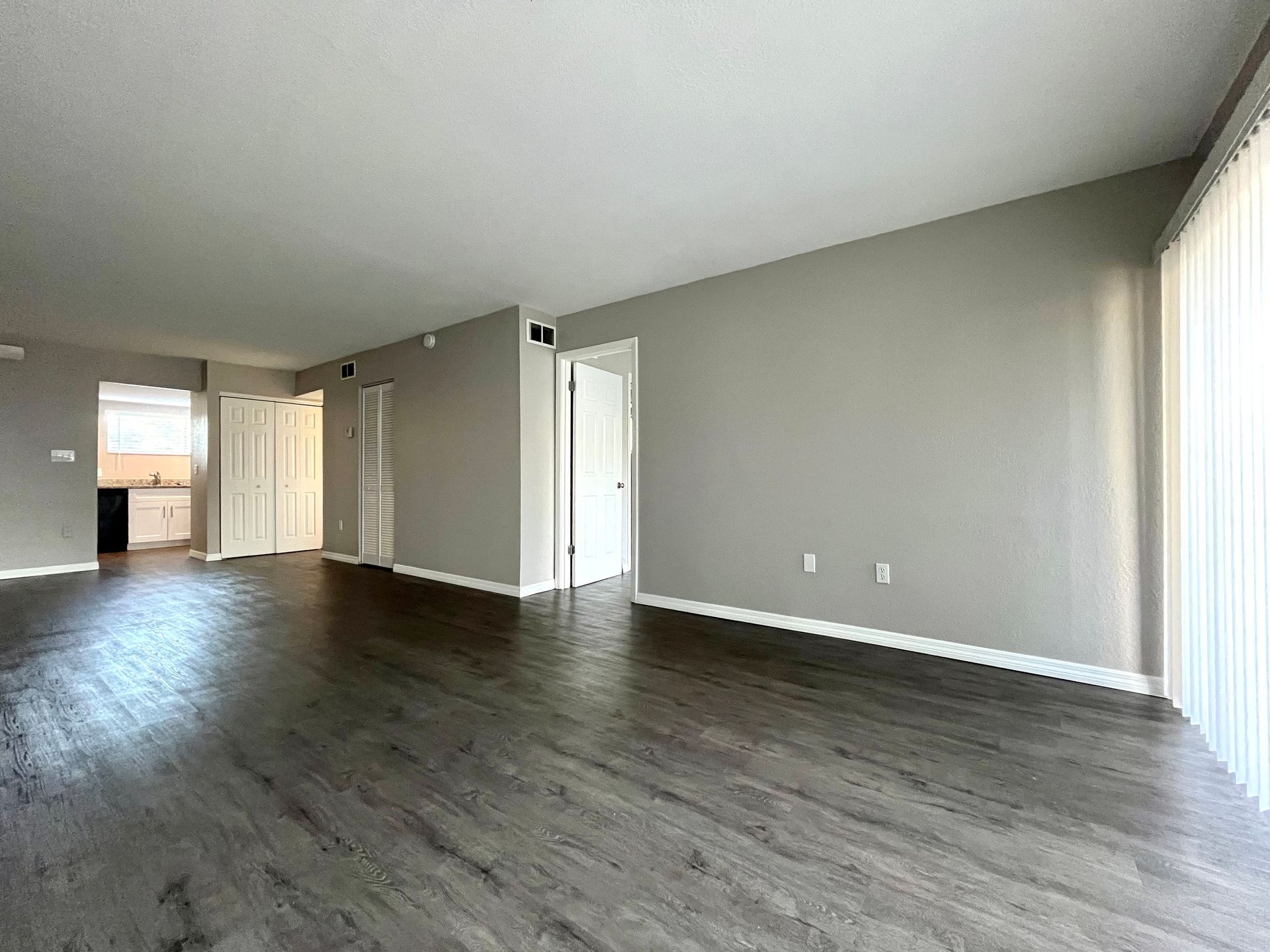 Empty living room with gray walls, dark wood-look floors, and vertical blinds.