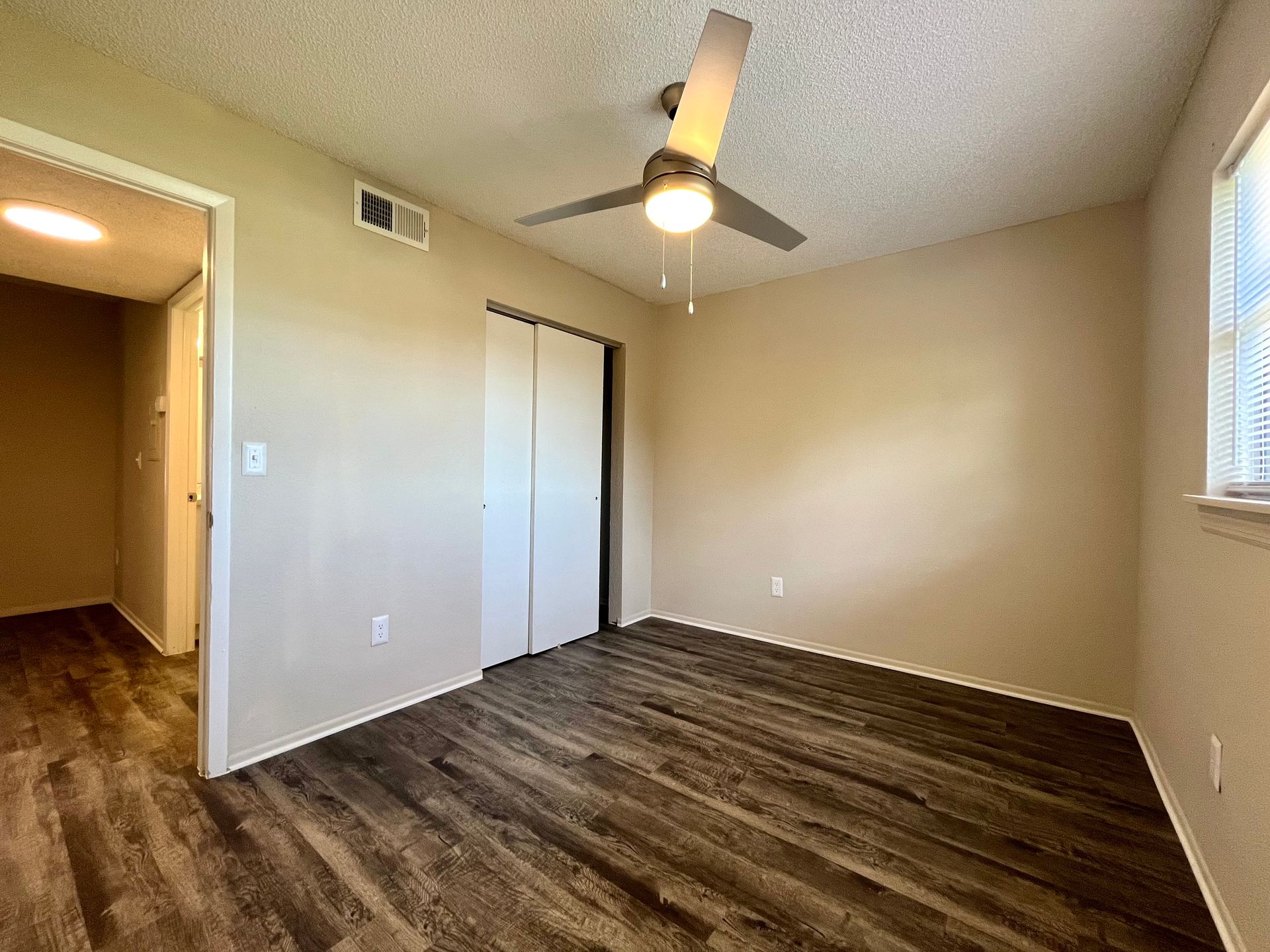 Bedroom with wood-look floor, beige walls, closet, door to hallway, and ceiling fan.