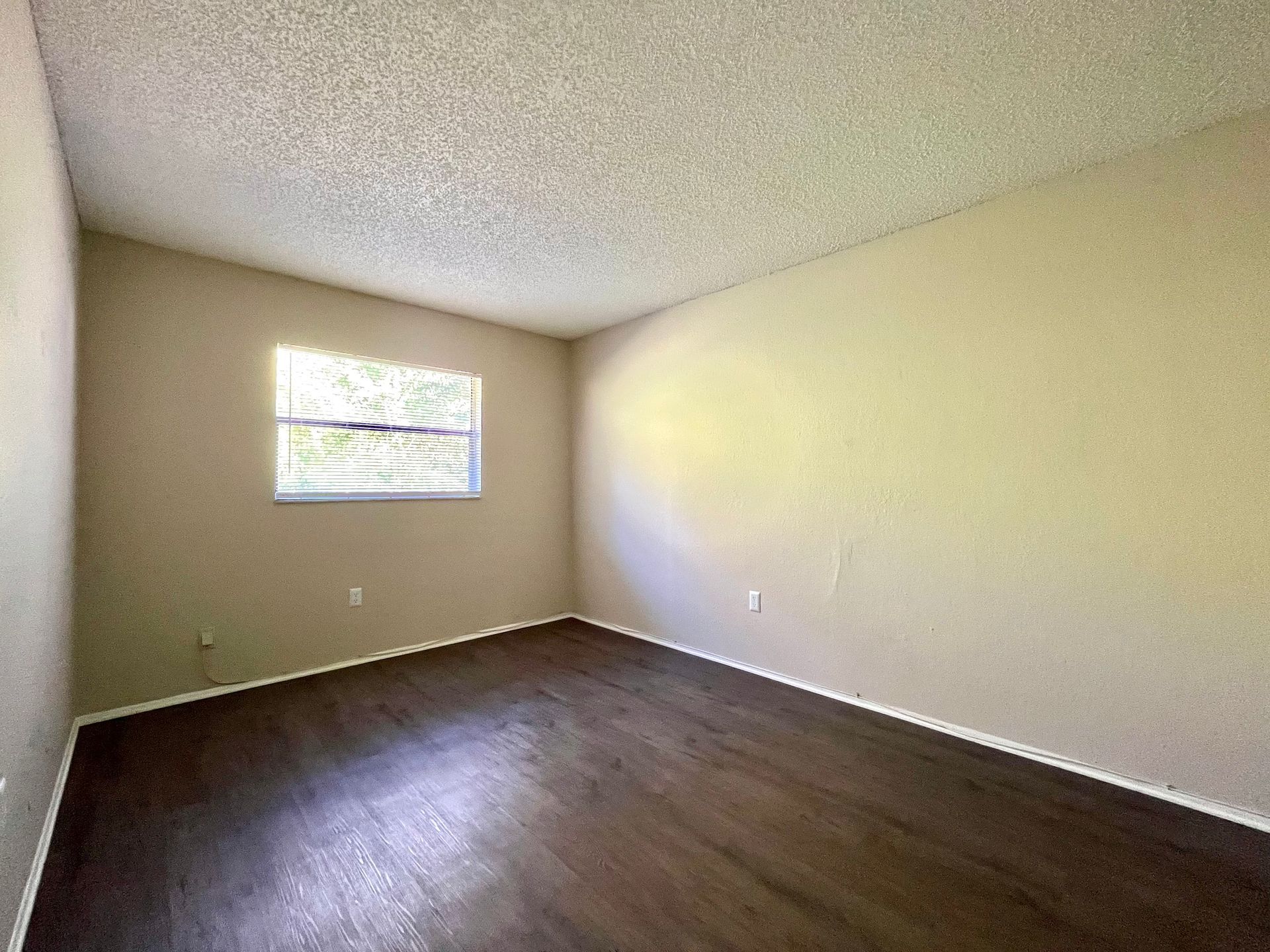 Empty bedroom with dark wood floor, tan walls, small window, and textured ceiling.