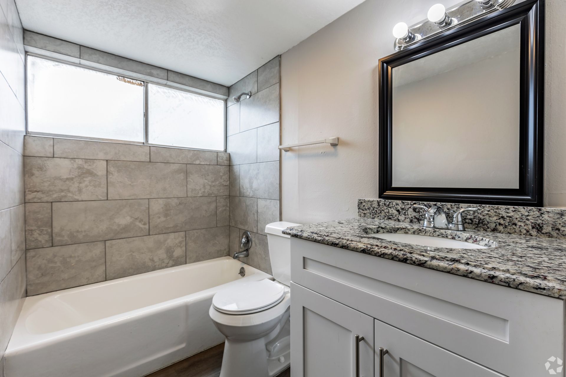 Bathroom with white vanity, gray countertop, tub, window with gray tile, and black-framed mirror.