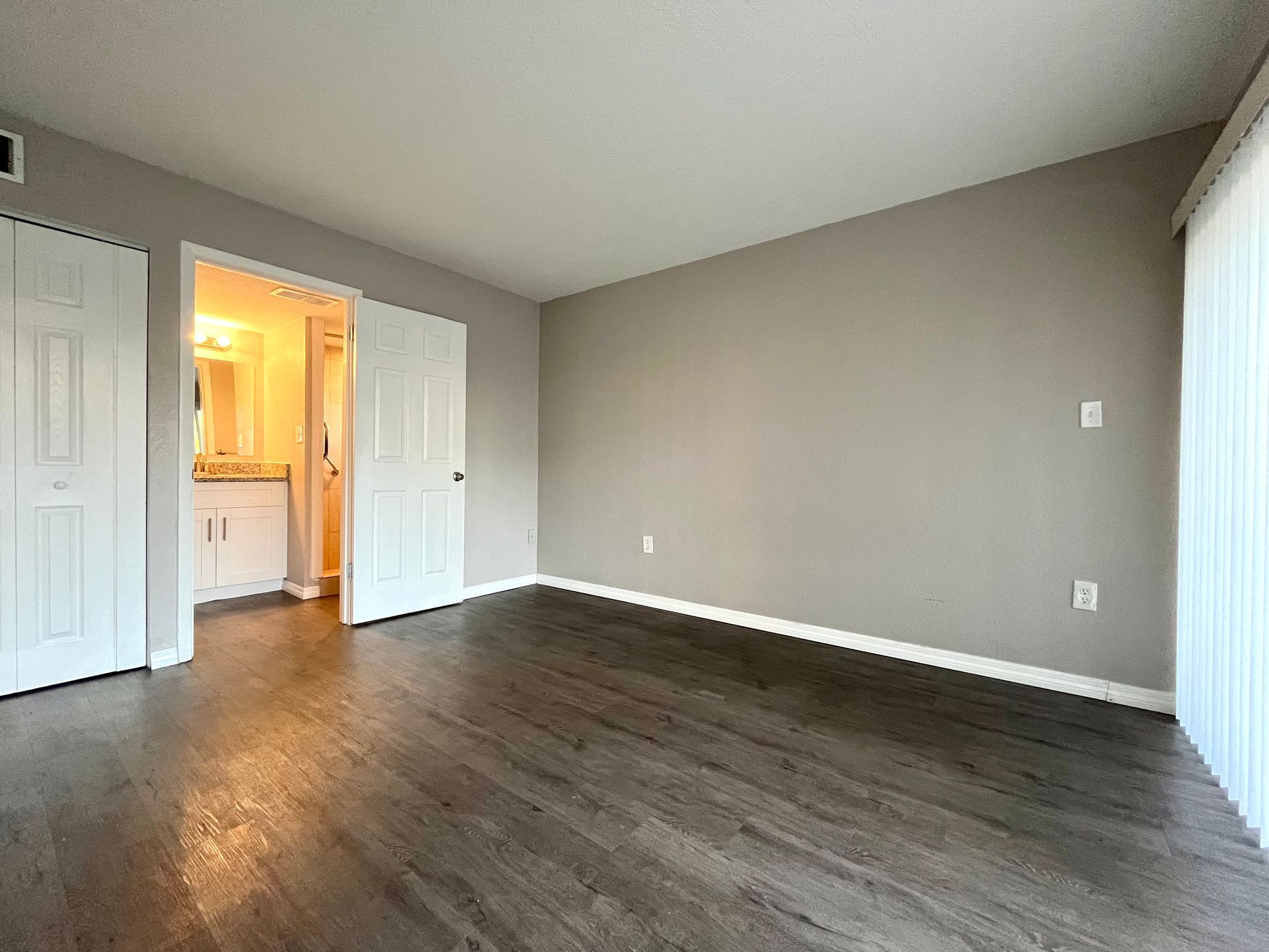 Empty bedroom with dark wood-look floors, gray walls, two white doors, and a doorway to a bathroom.