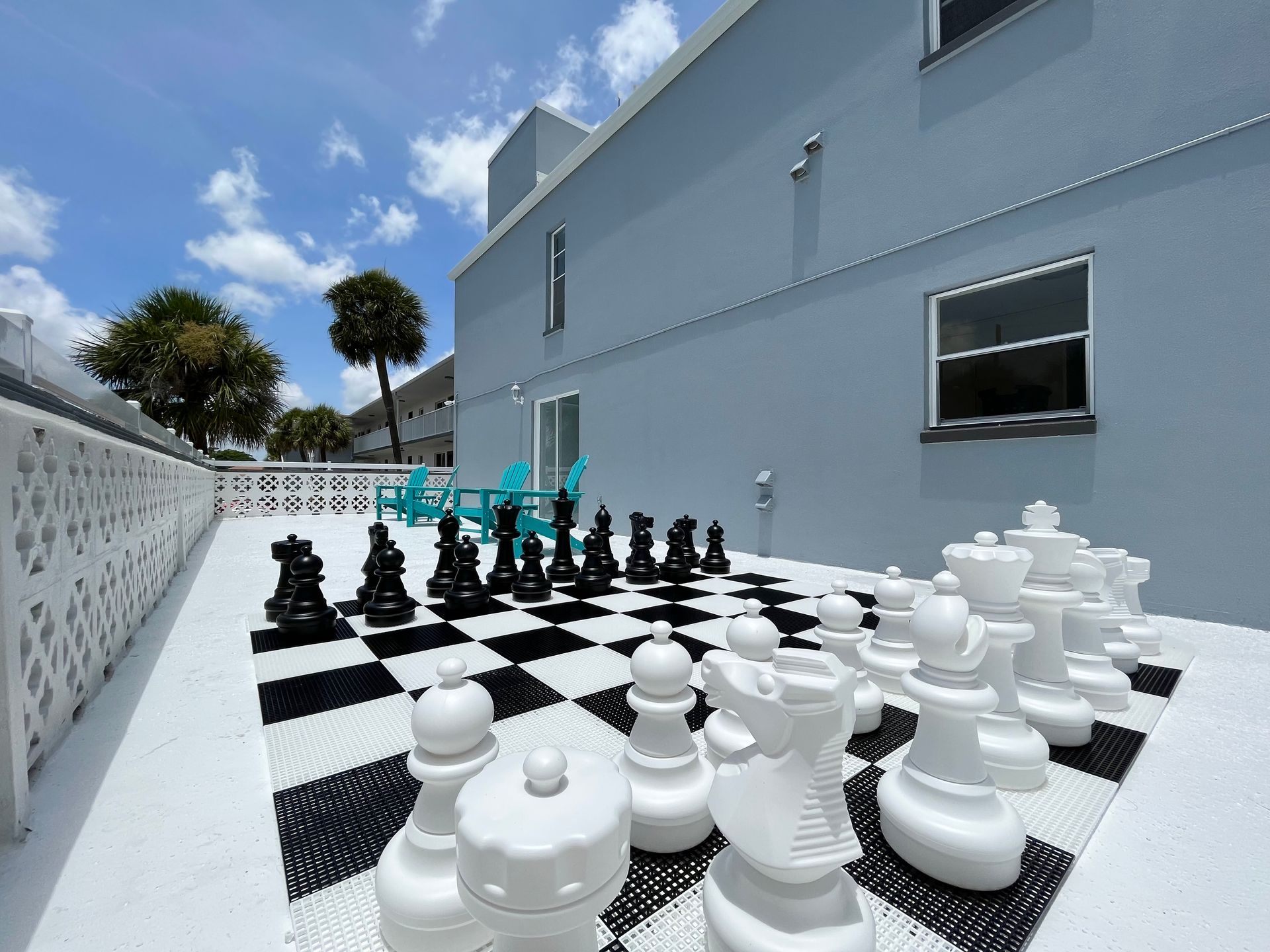 Giant chess set on a checkered mat on a patio, overlooking a blue building and sky.
