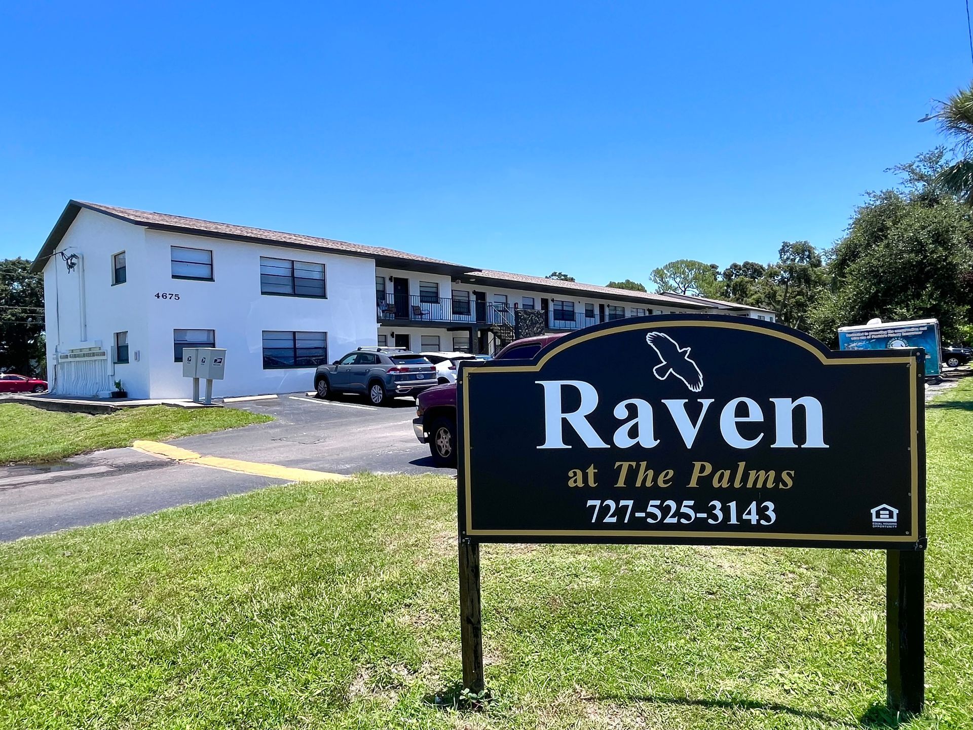 Raven at the Palms apartment complex, sign in foreground, two-story white building in the background, blue sky.