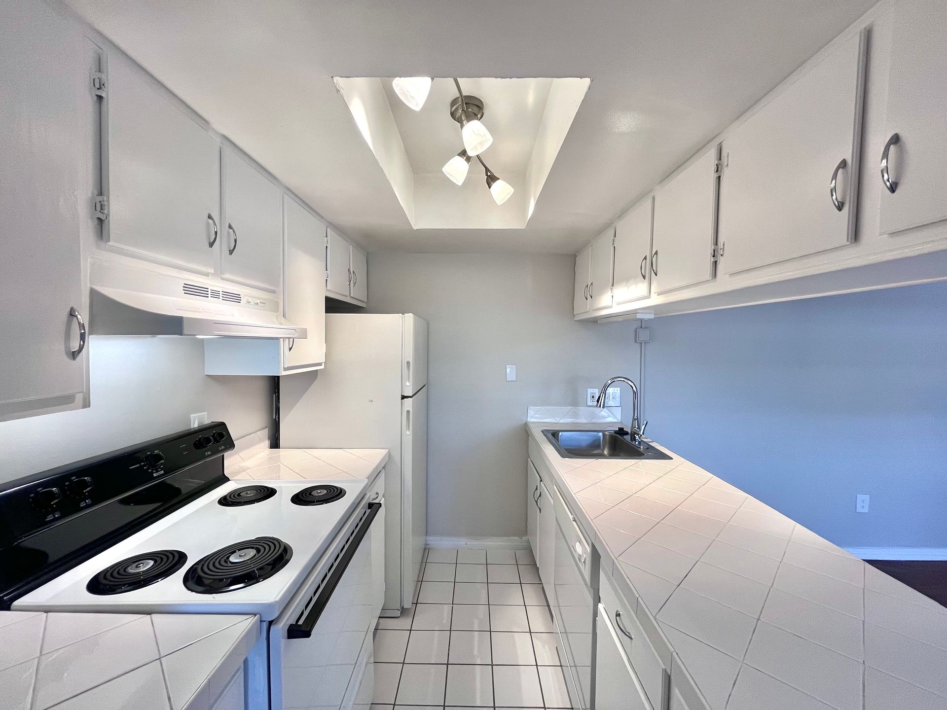 Narrow, white kitchen with overhead cabinets, stove, refrigerator, and sink.