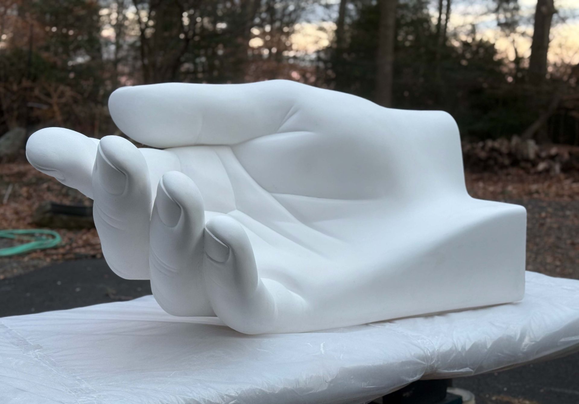 Large white hand sculpture, palm up, sitting on a white surface. Outdoors, trees in the blurred background.