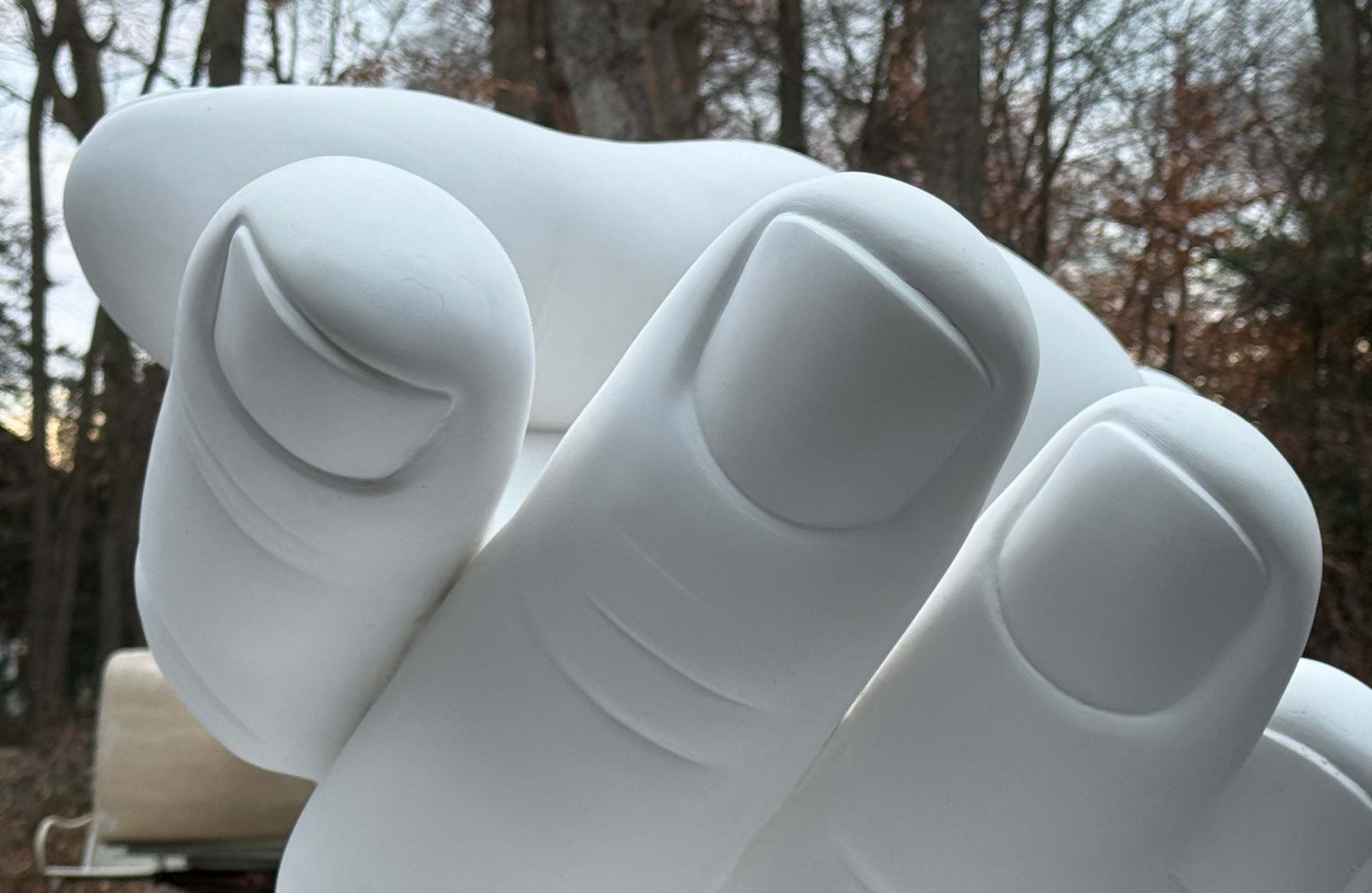 White hand sculpture reaching forward, outdoors with trees in background.