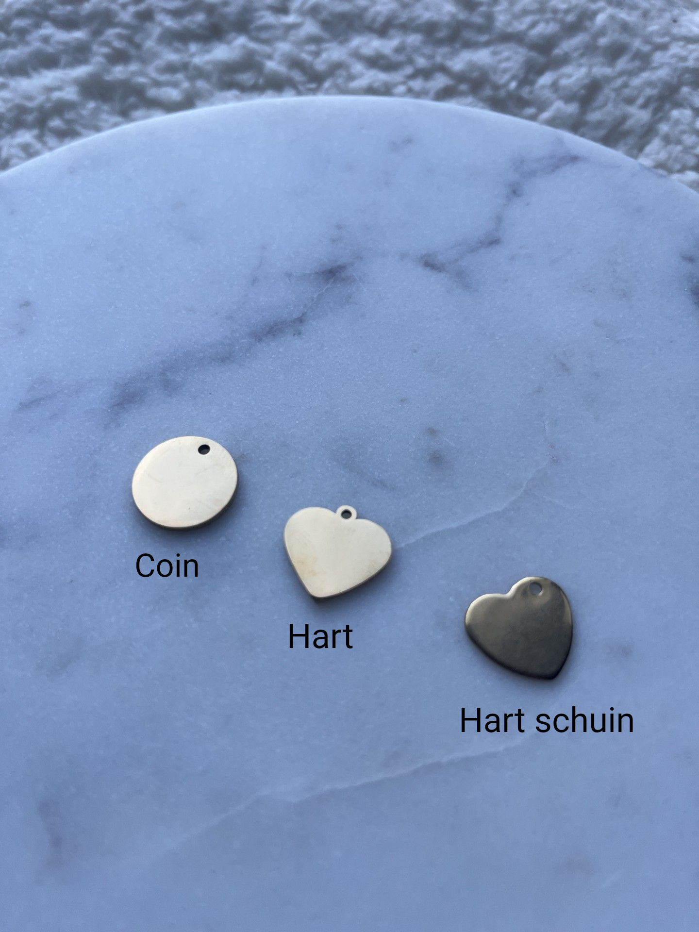 Verschillende gepersonaliseerde kettingen met hart- en ronde hangers liggen uitgestald op een marmeren ondergrond naast een klein mauve fluwelen zakje.