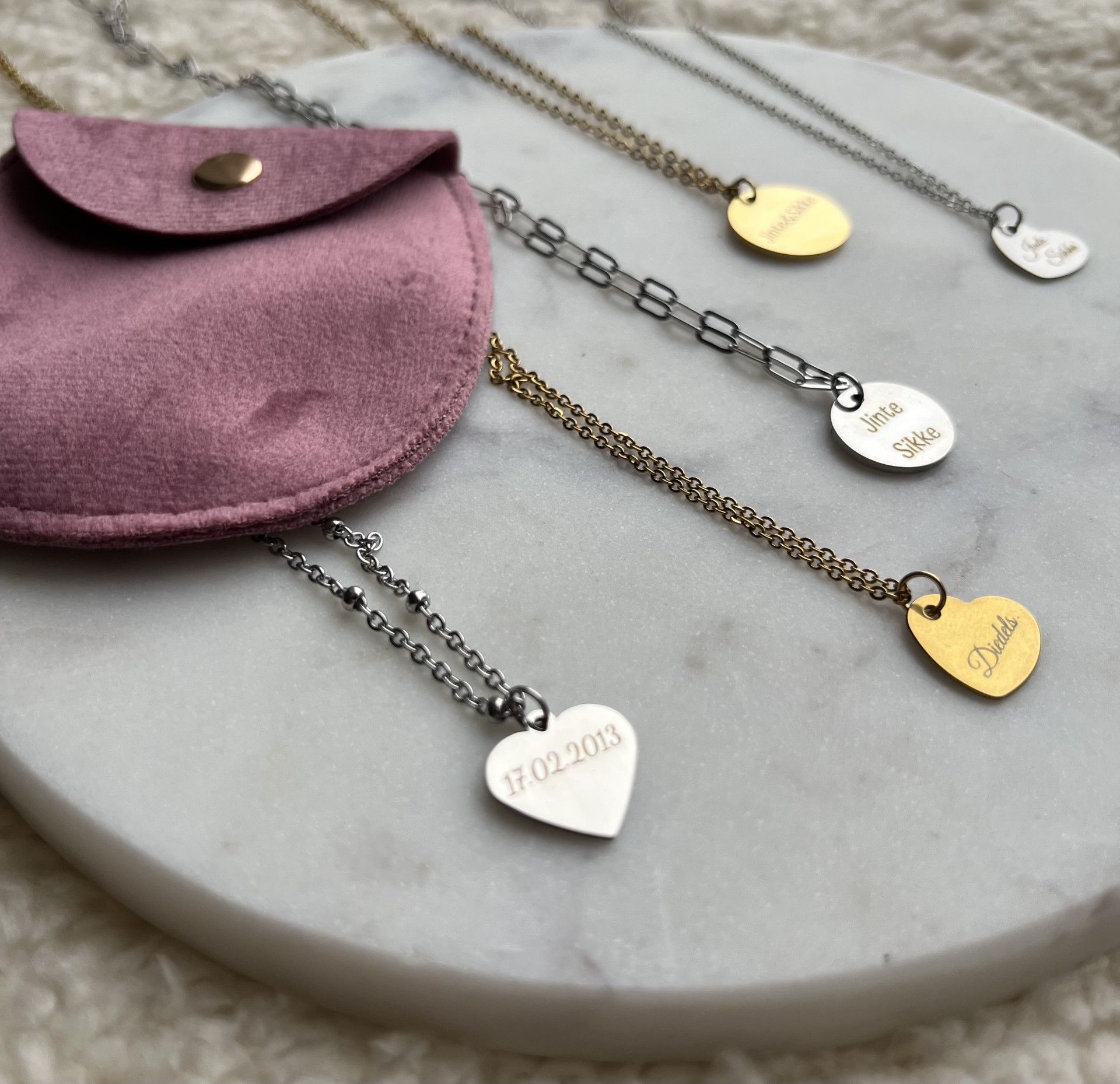 Verschillende gepersonaliseerde kettingen met hart- en ronde hangers liggen uitgestald op een marmeren ondergrond naast een klein mauve fluwelen zakje.