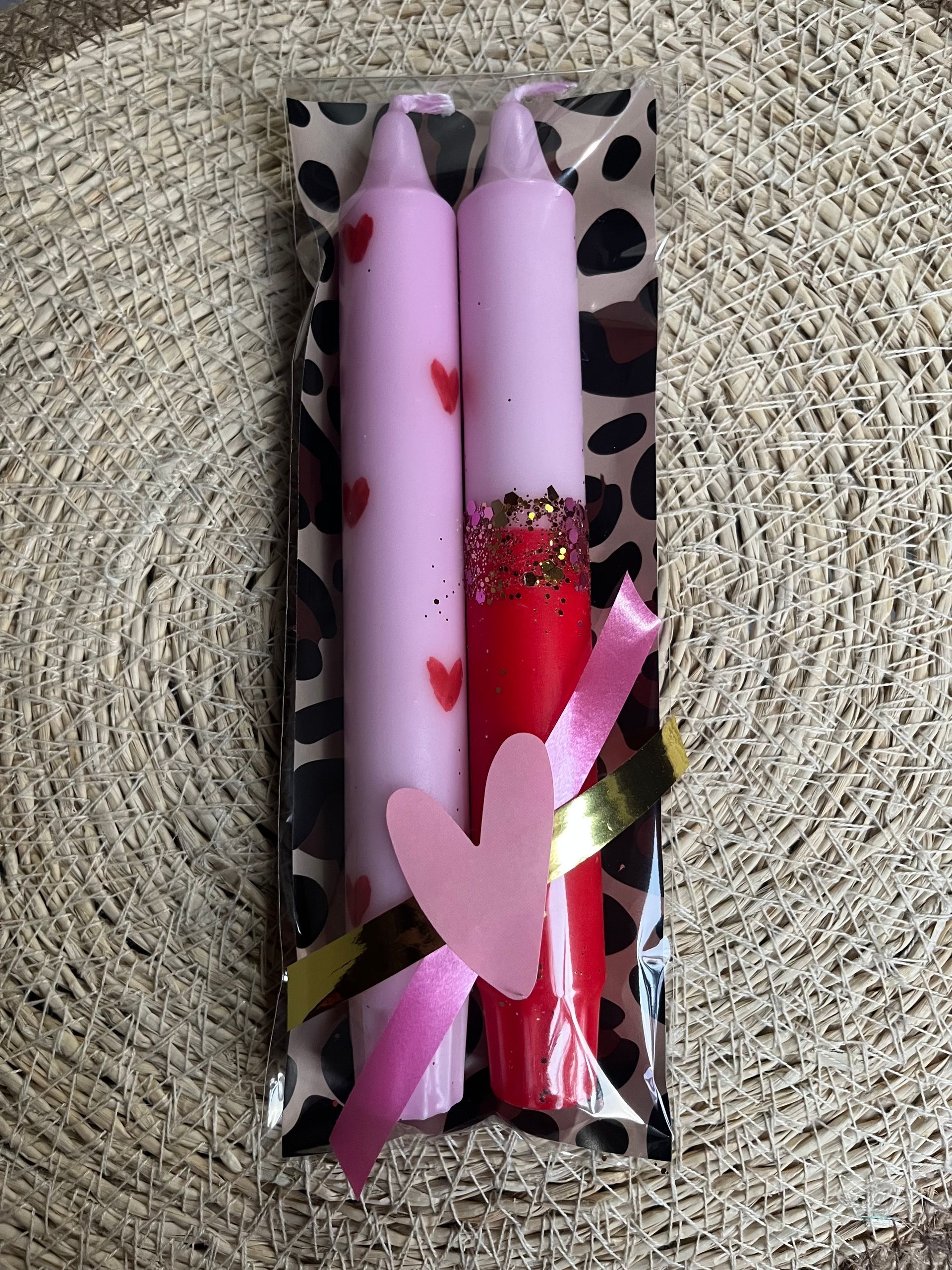 Twee taps toelopende kaarsen – een lichtroze met rode hartjes en een halfroze, halfrode met glitter – verpakt met een hartvormig label.