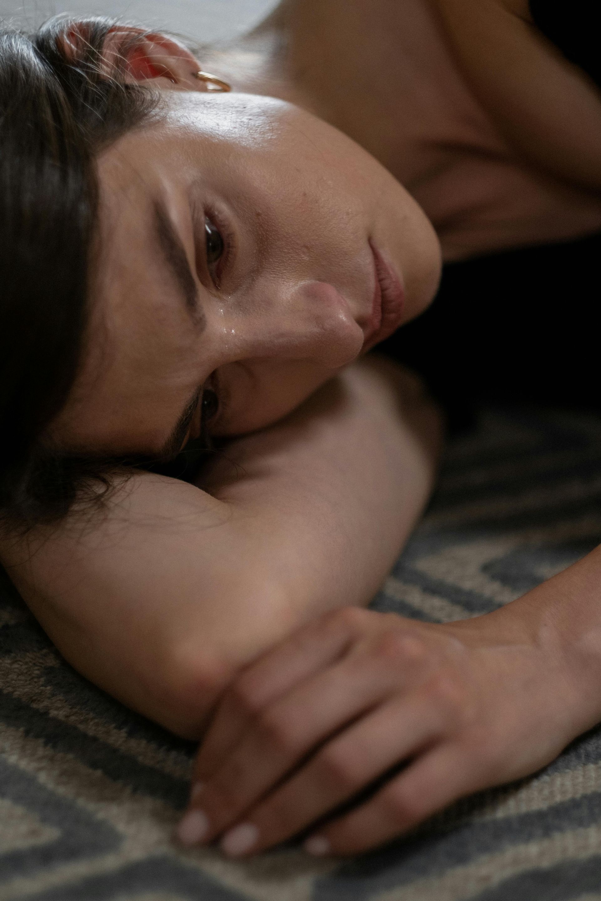 Person lying on patterned carpet, head resting on arms, looking down, somber expression.