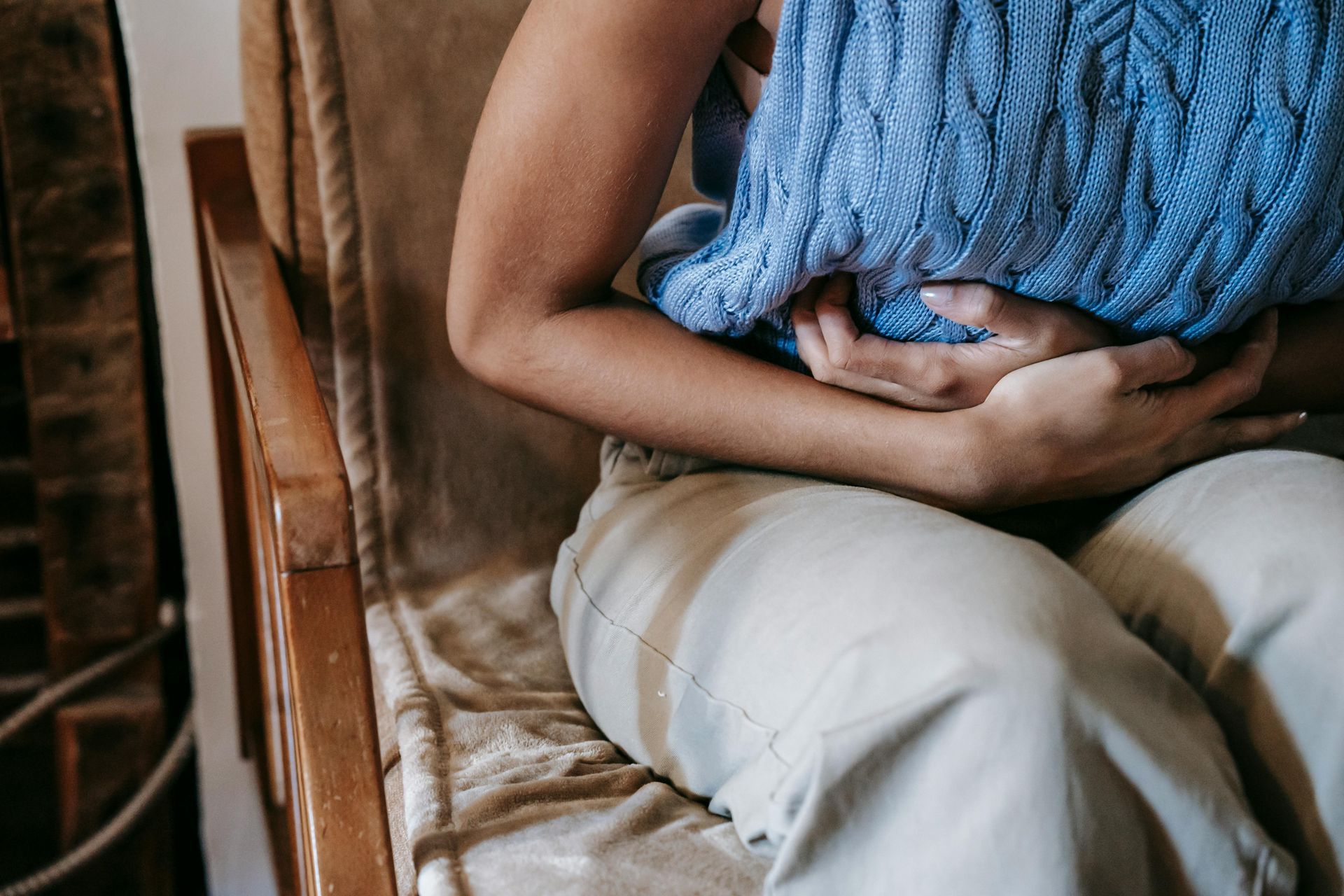 Person seated, clutching stomach in discomfort. Wearing blue sweater, beige pants; sitting in a chair.