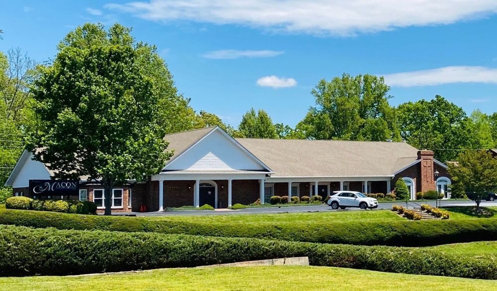Low brick building with white trim, tan roof, green bushes, blue sky. Car parked out front.