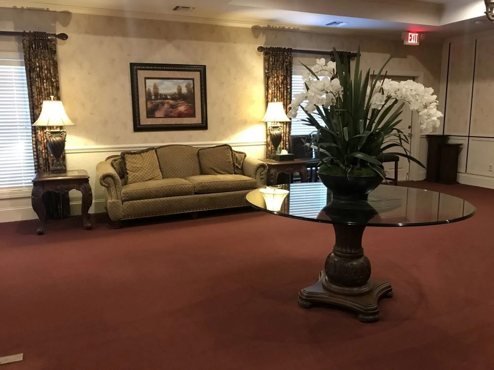 A waiting room with red carpet, couch, lamps, round table with flowers, and painting.