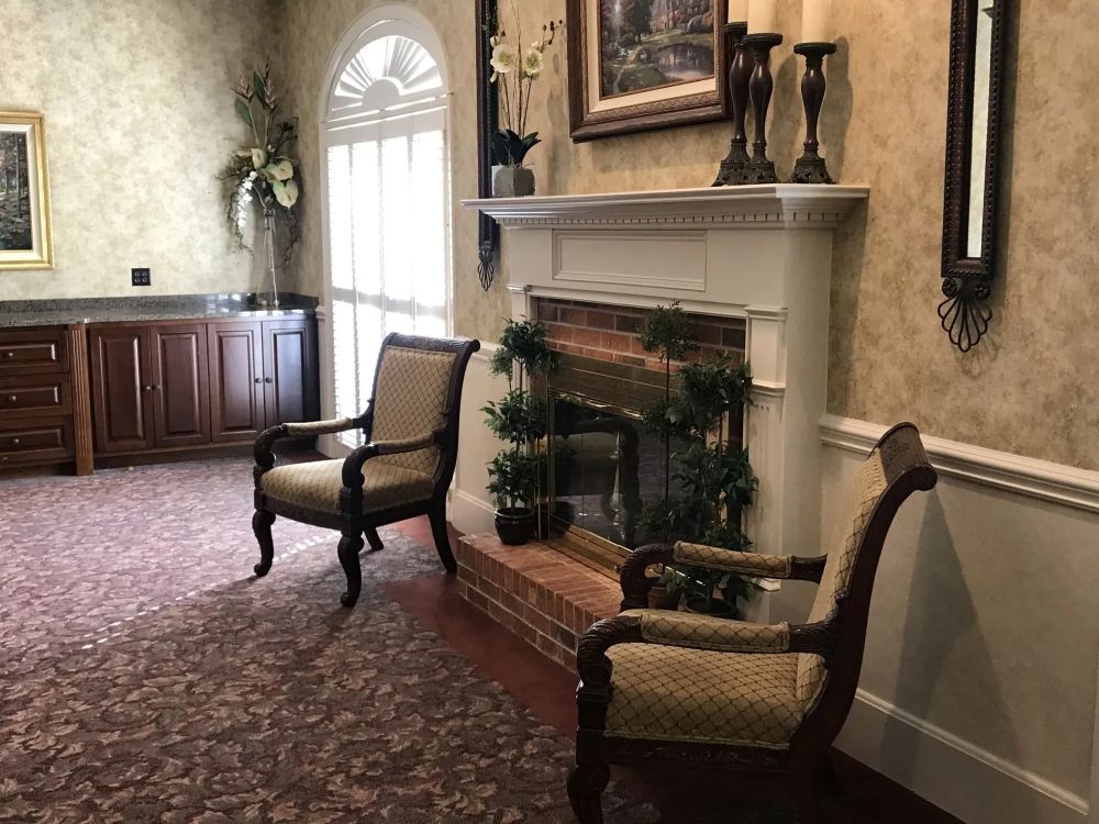 Two ornate chairs face a brick fireplace with greenery. Wooden cabinetry and floral wallpaper.