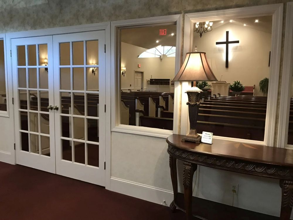 Interior view of a church through a window, showing pews, a cross, and a lamp on a table.