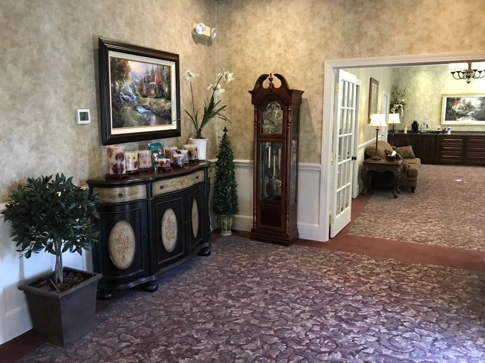 Hallway with ornate furniture, painting, grandfather clock, and carpet. Doorway leads to another room.