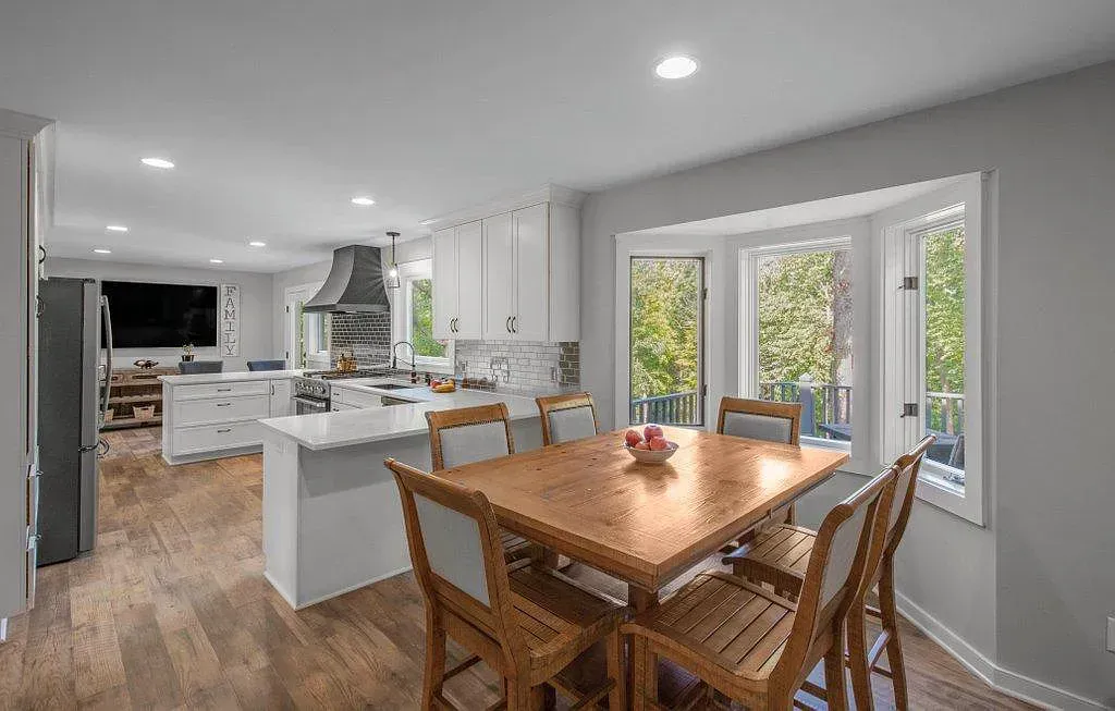 Bright open kitchen and dining area with wood table, white island, and large windows overlooking greenery
