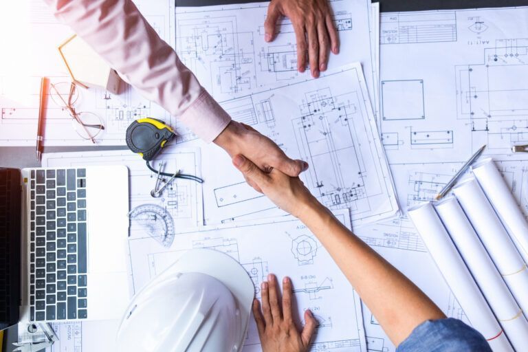 Hands meeting over architectural blueprints beside a laptop and drafting tools