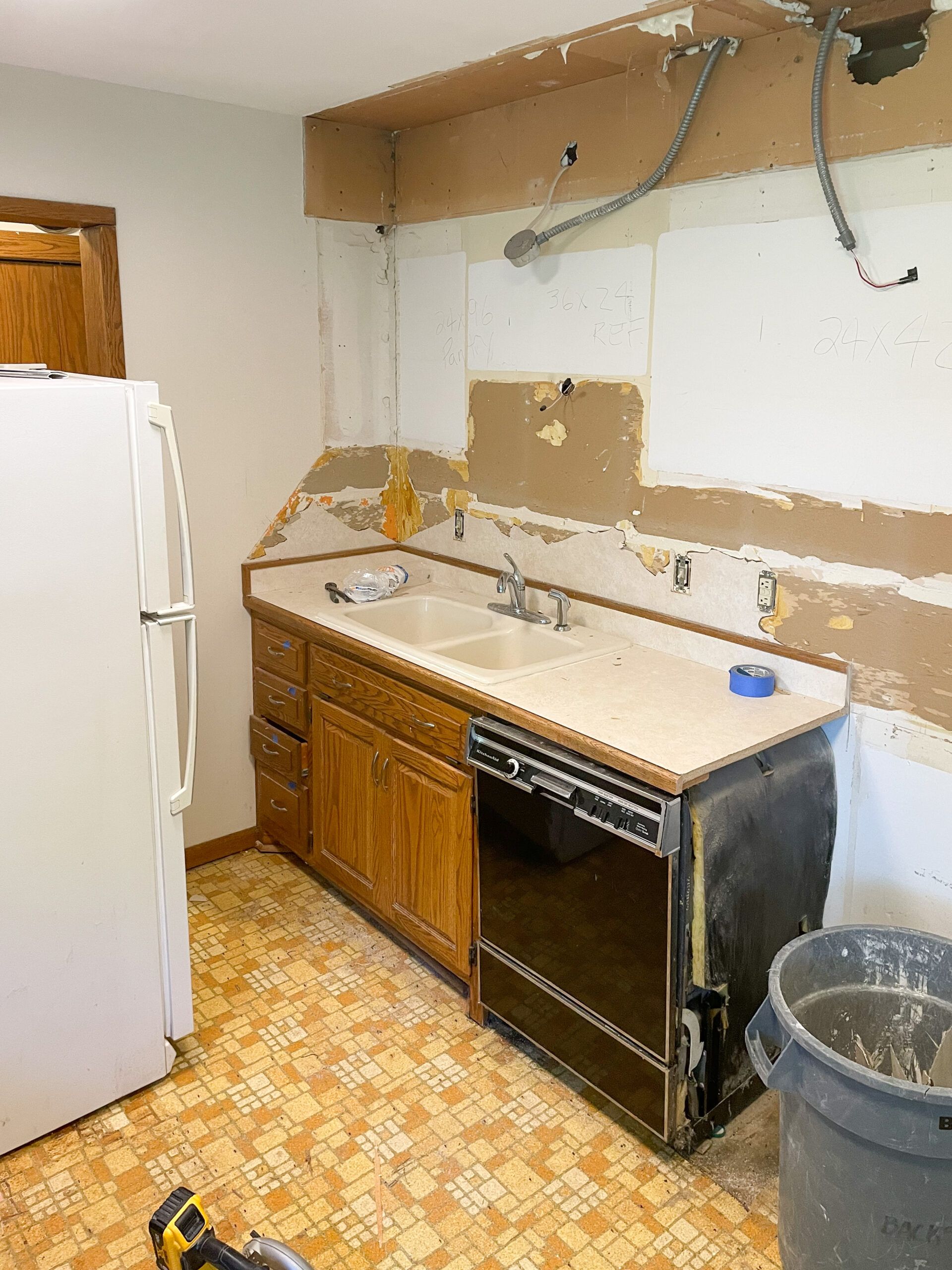 Damaged kitchen with a sink, missing cabinets, exposed wall, refrigerator, and trash can