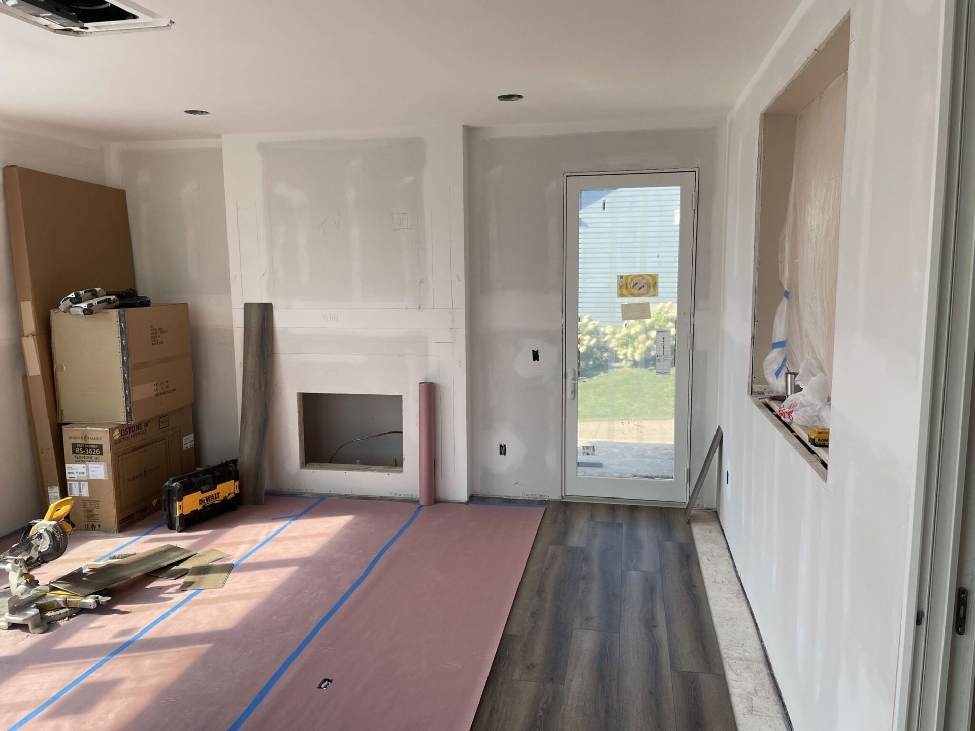Renovation room with drywall, taped floor, a fireplace, stacked boxes, and a glass door to outside. Renovation room with drywall, taped floor, a fireplace, stacked boxes, and a glass door to outside.