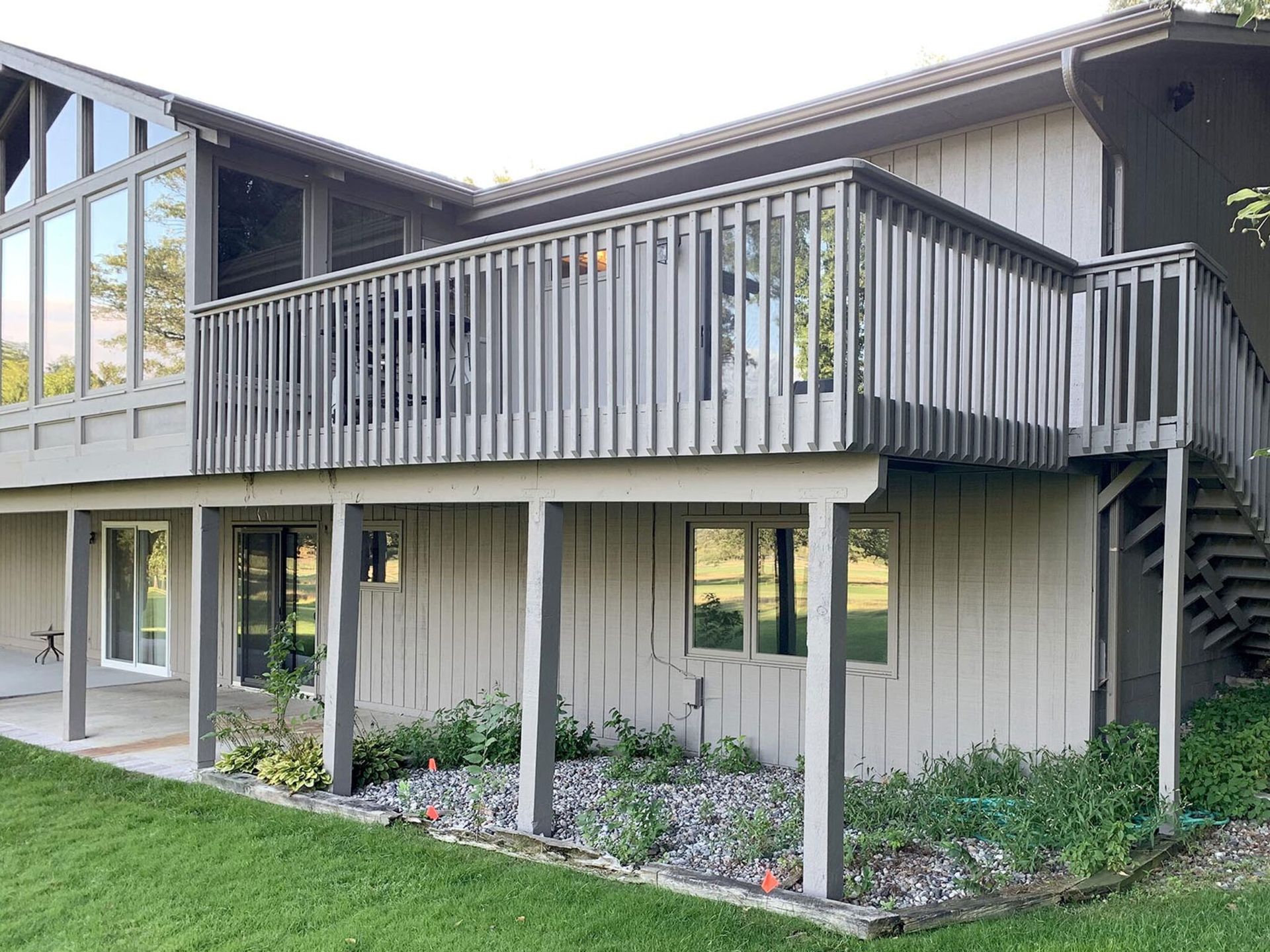 Two-story beige house with gray balcony and stairs above a green lawn and garden beds Two-story beige house with gray balcony and stairs above a green lawn and garden beds