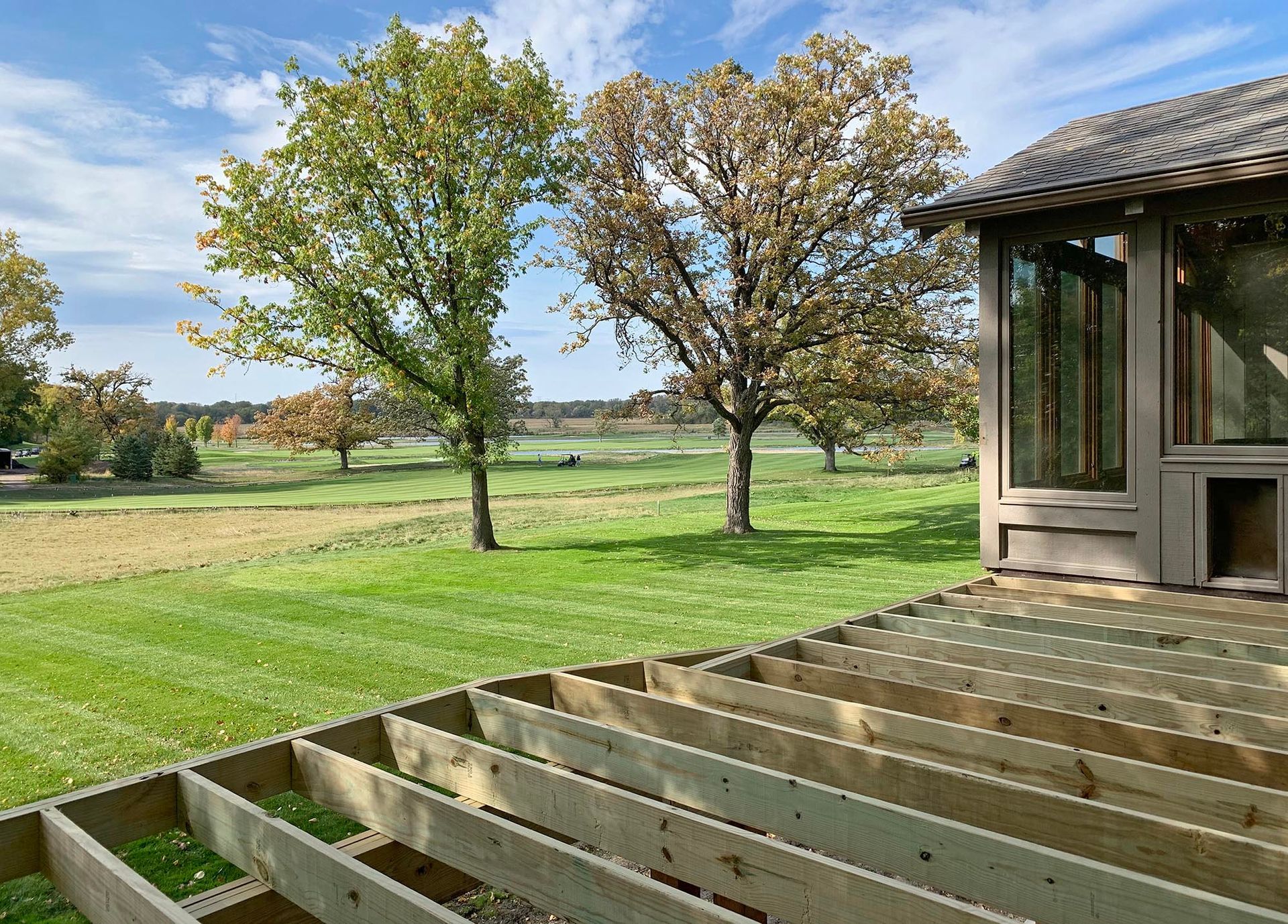 Wooden porch overlooking a green field with two trees and a small rustic building under a blue sky