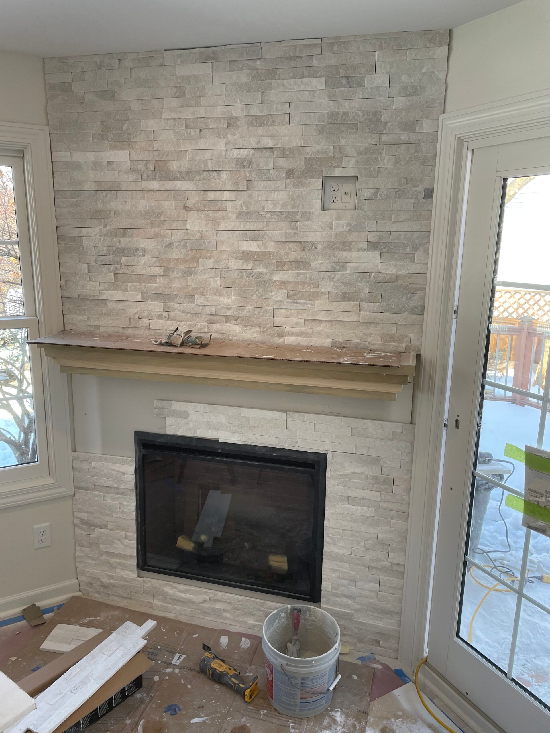 Empty living room with stone fireplace, wooden mantel, and glass door letting in daylight.