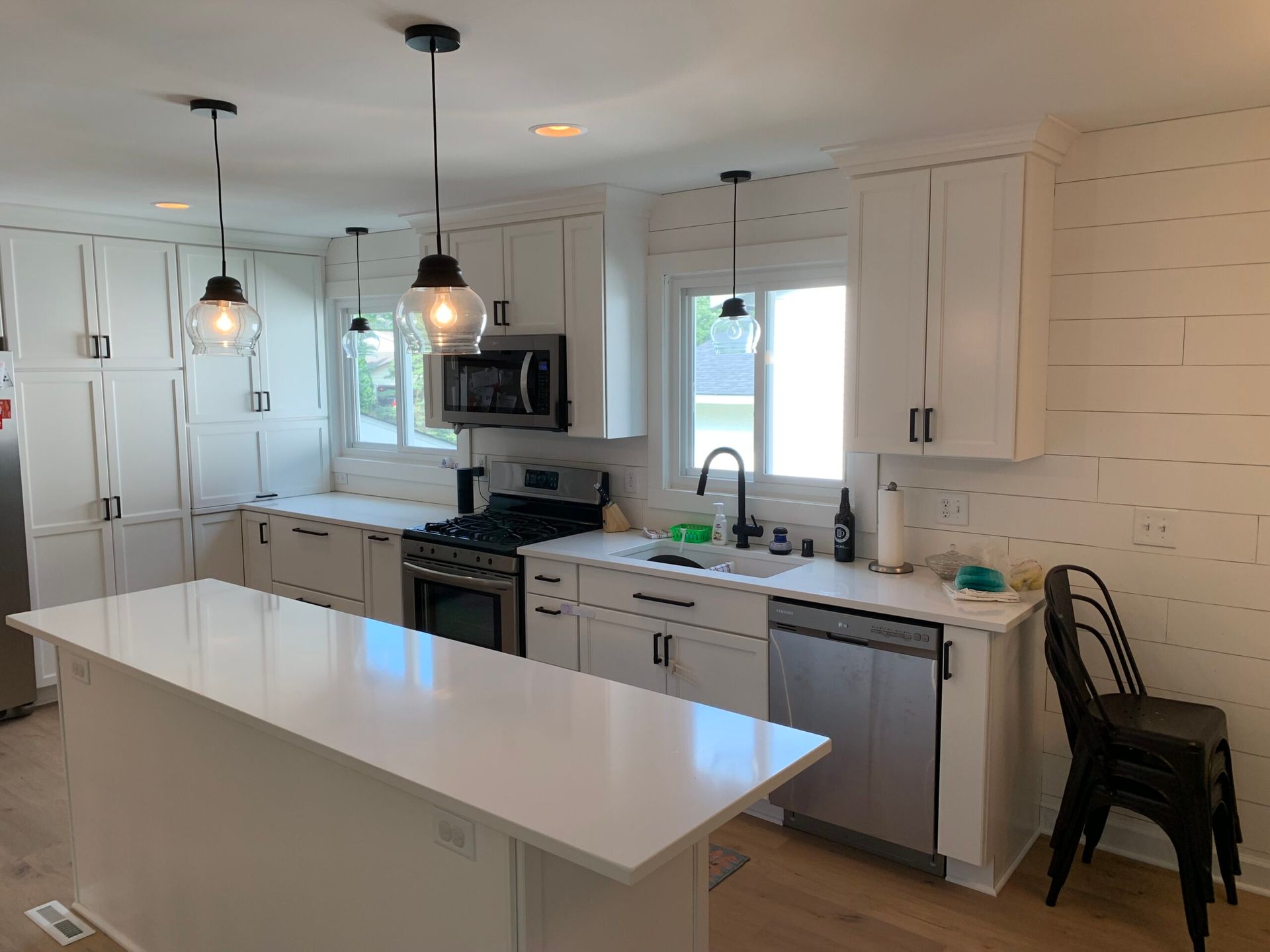 Bright white kitchen with island, stainless appliances, pendant lights, and stack of chairs by the wall