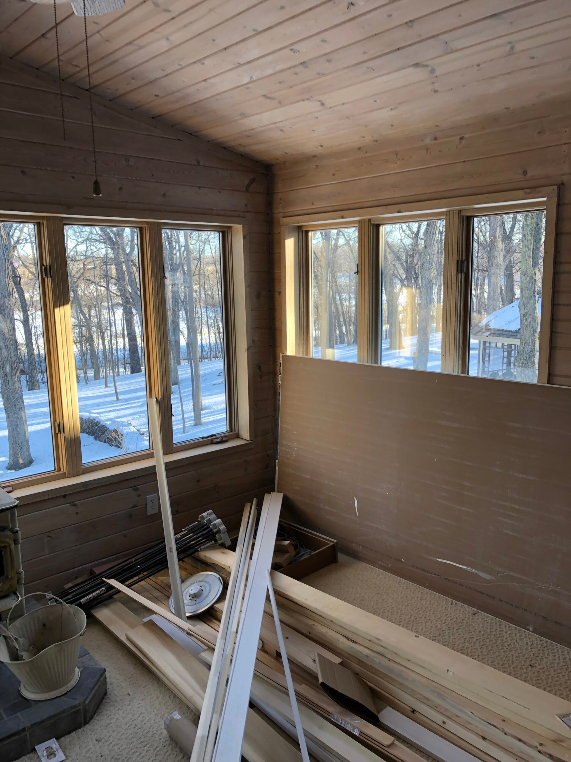 Sunlit unfinished room with large windows, wood walls and ceiling, and stacks of boards on the floor