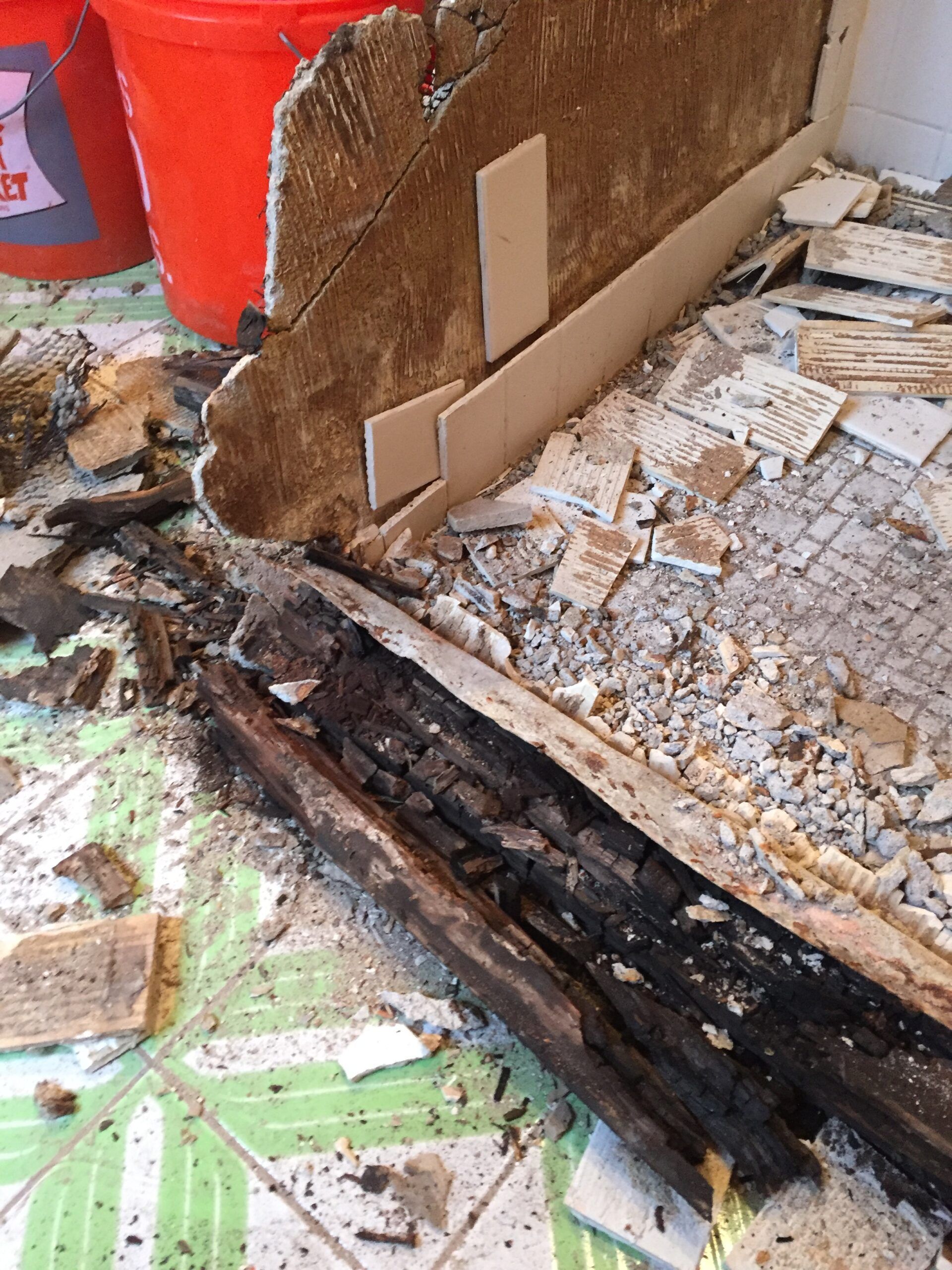 Water-damaged floor with blackened, rotting wood and debris beside a red wall.