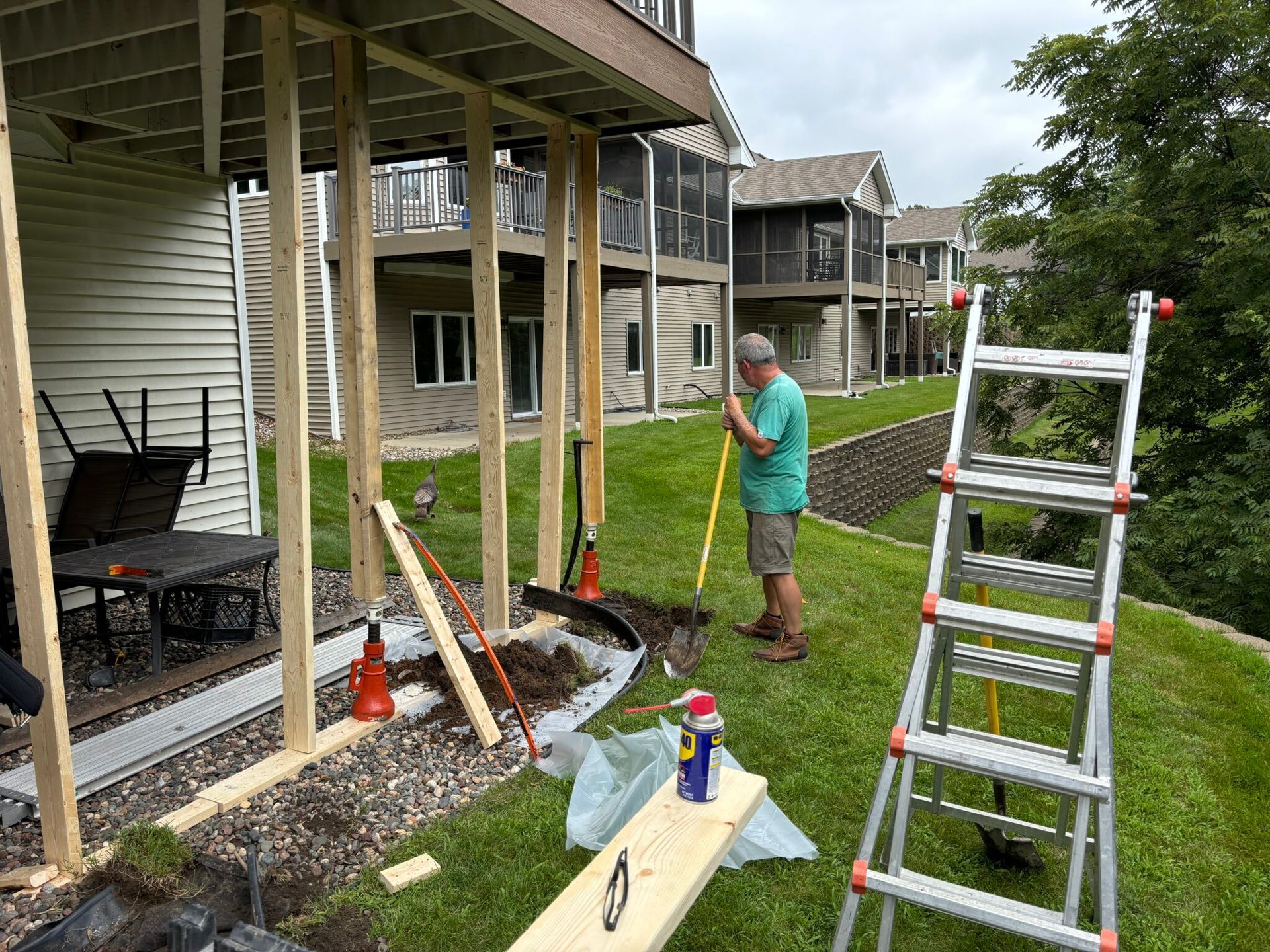Person working on landscaping beside a house, with tools, gravel, and a ladder on the lawn.