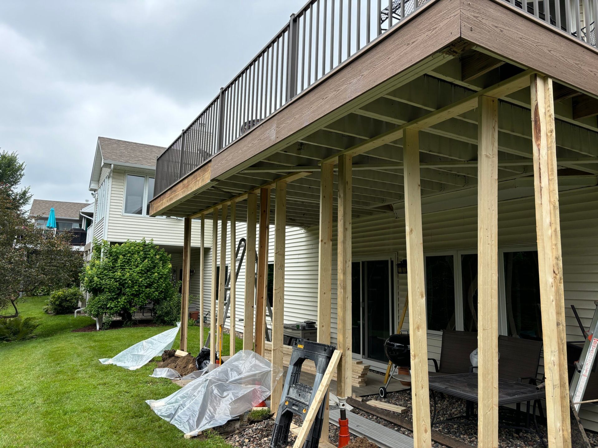 Raised deck under construction beside a house, with wooden posts, railing, and building materials on grass.