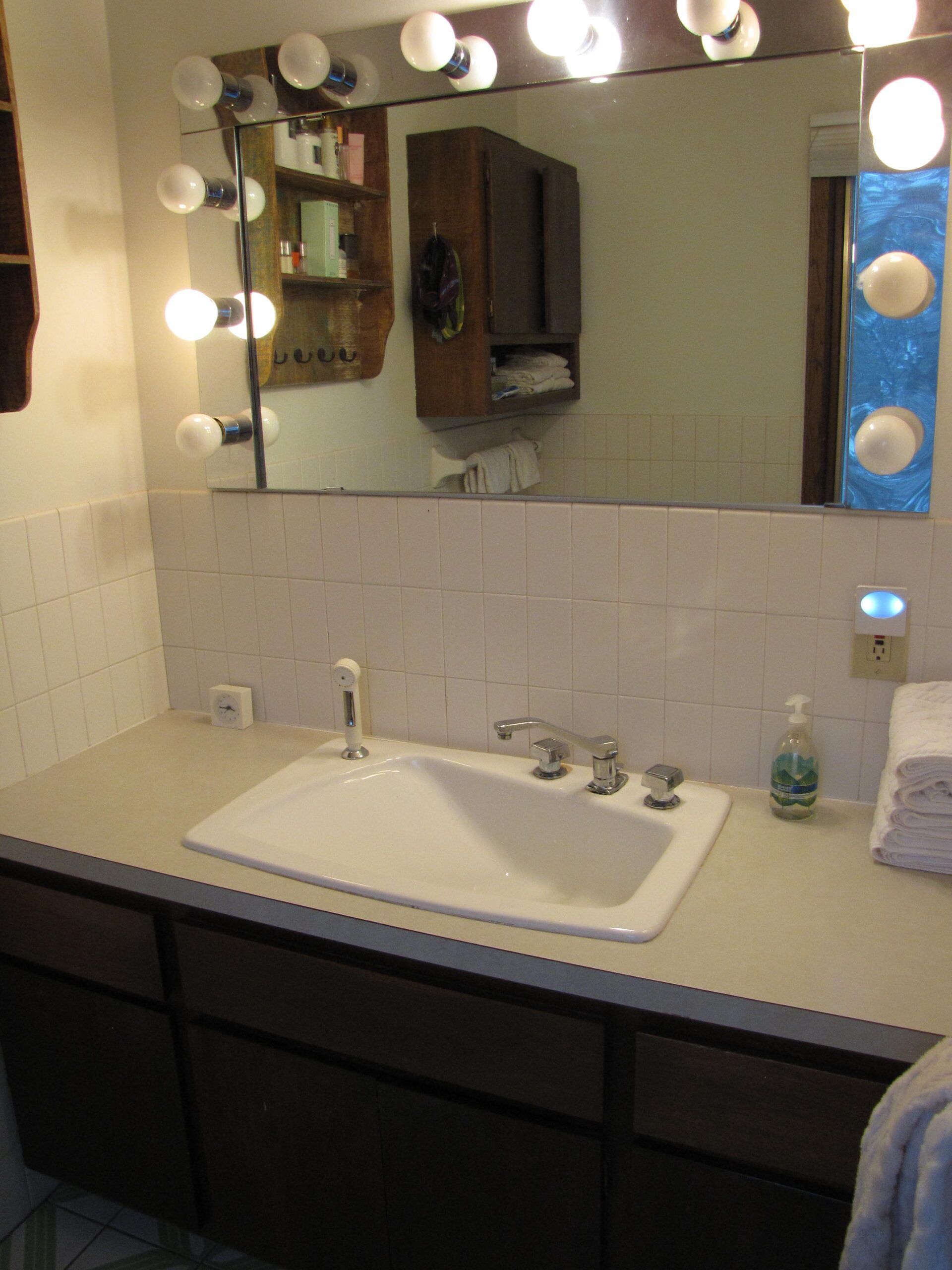 Bathroom vanity with large mirror, lit bulbs, and white sink on a light countertop.