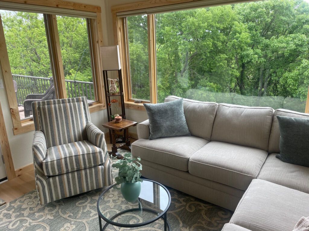 Bright sunroom with gray sectional, striped armchair, glass table, and lush green trees outside.
