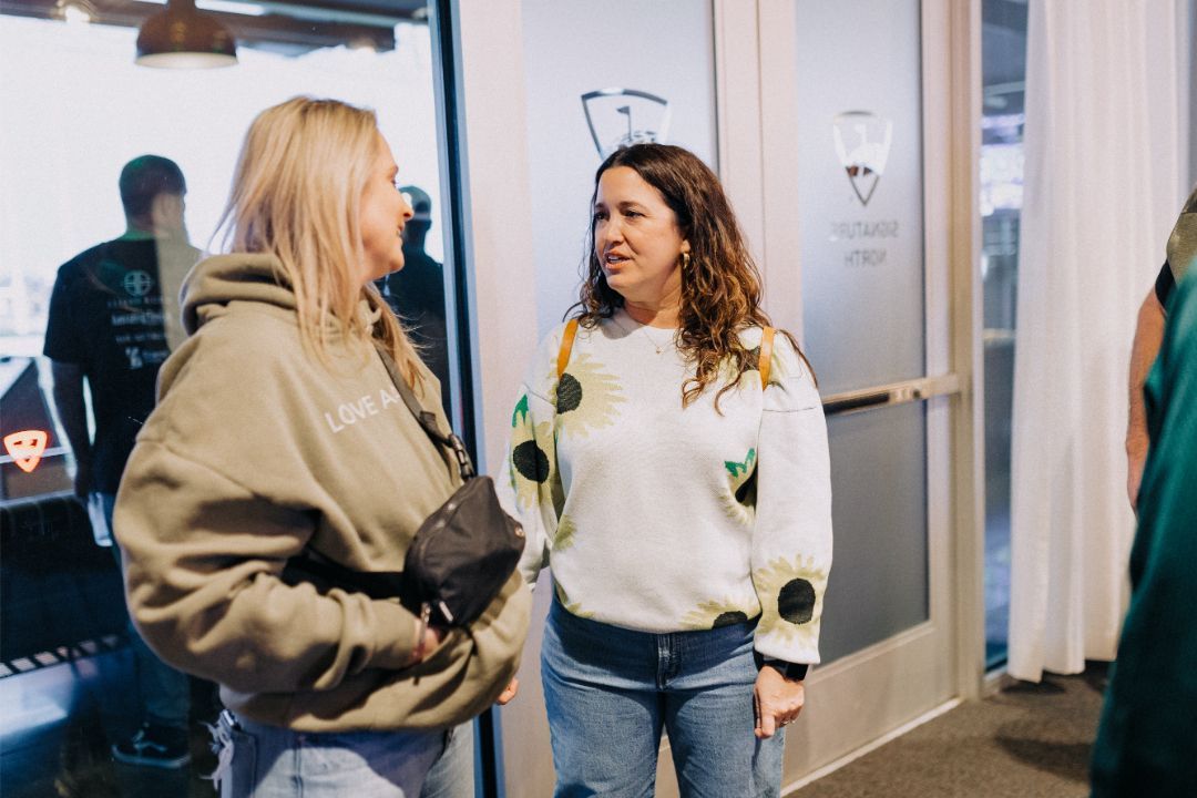 Two women are standing next to each other in a hallway talking to each other.