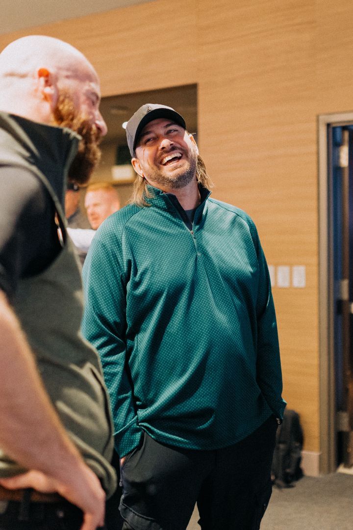 A man in a green sweater is laughing while standing next to another man.