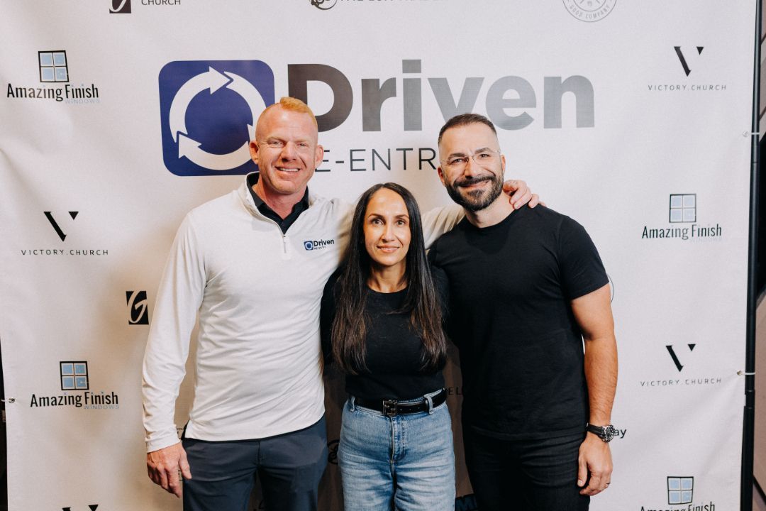 Three people are posing for a picture in front of a sign that says driven.