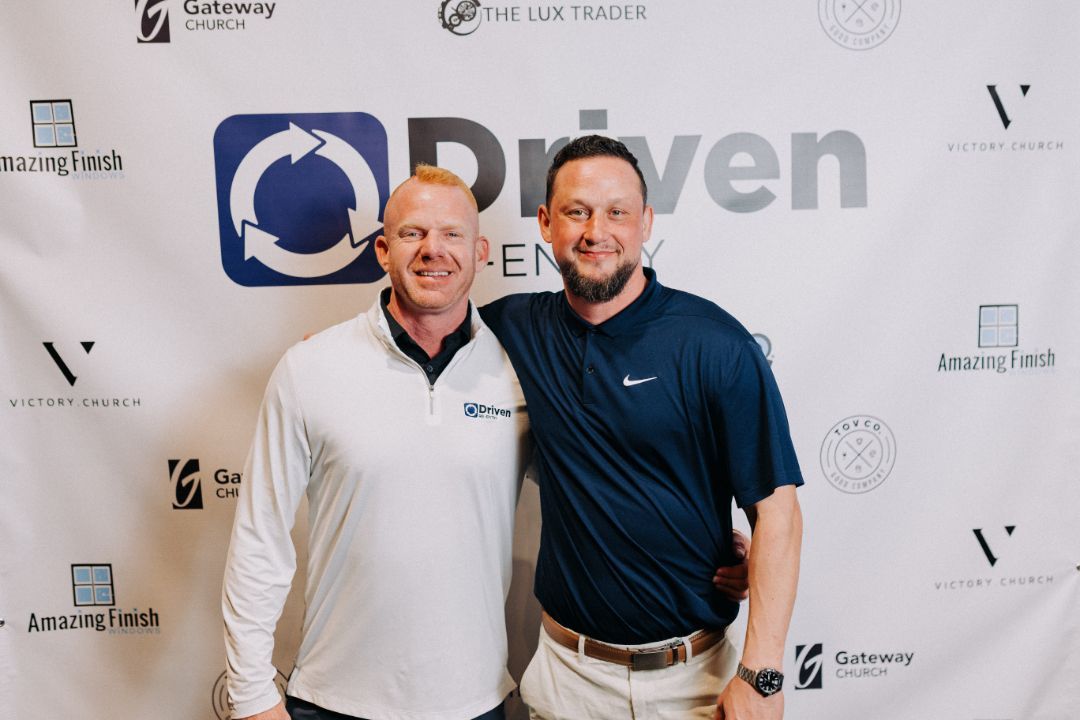 Two men are posing for a picture in front of a driven banner.