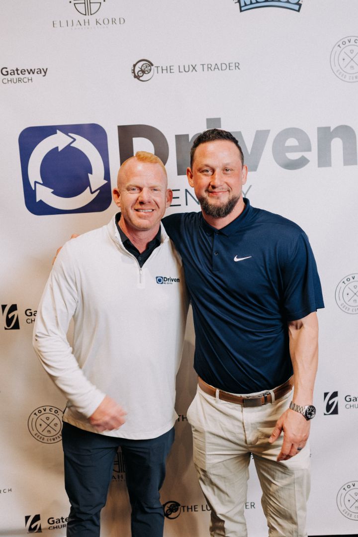 Two men are posing for a picture in front of a sign that says driven.