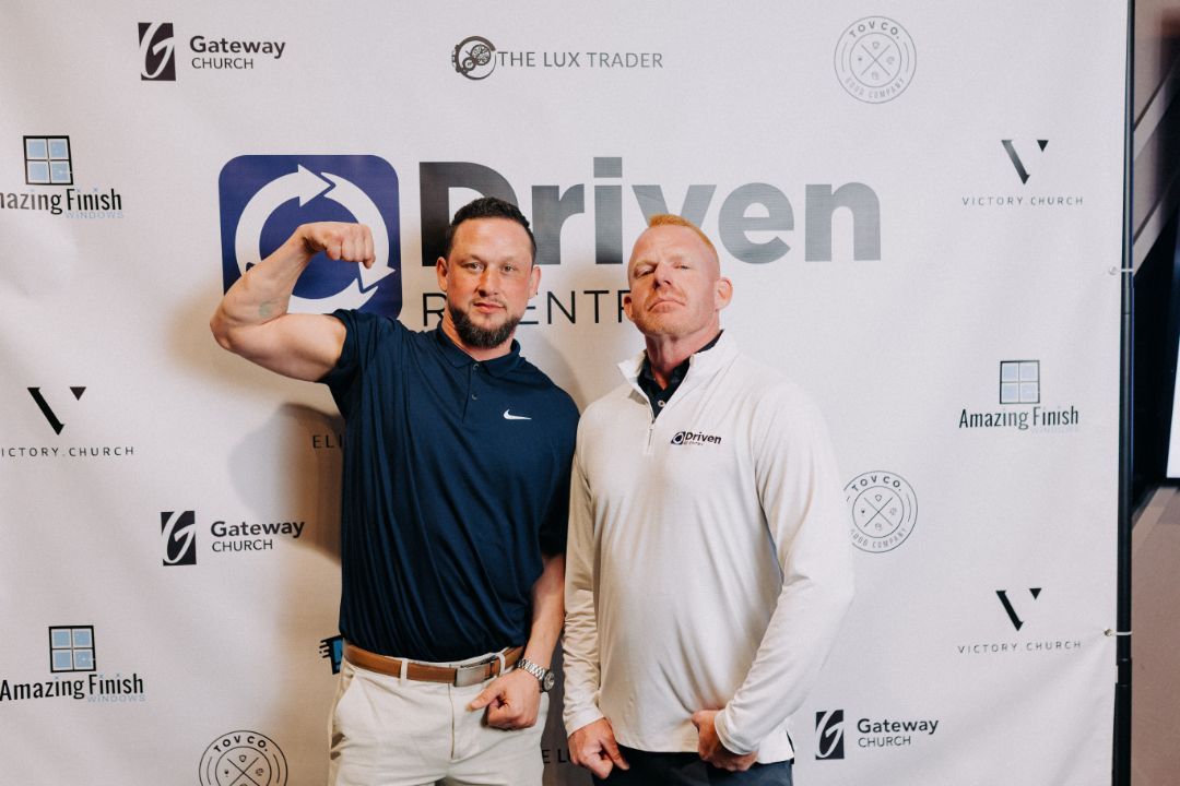 Two men are posing for a picture in front of a sign that says driven.