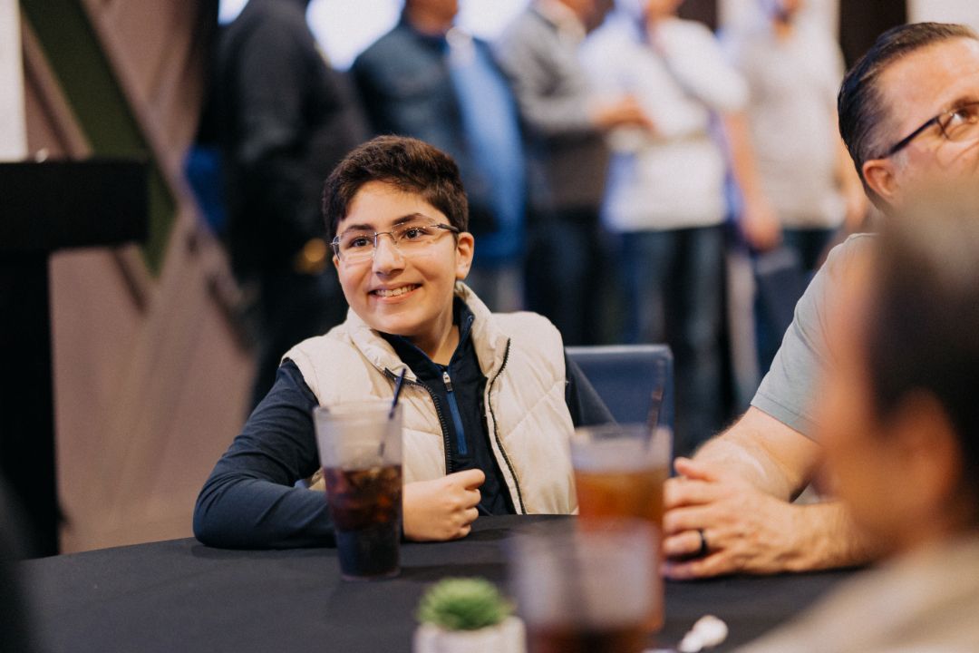 A young boy is sitting at a table with a drink in his hand.