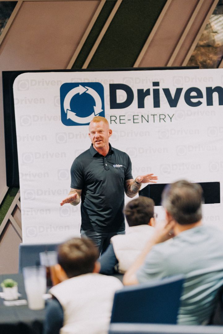 A man is giving a presentation in front of a sign that says driven