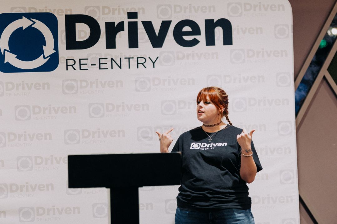 A woman is standing at a podium in front of a sign that says driven re-entry