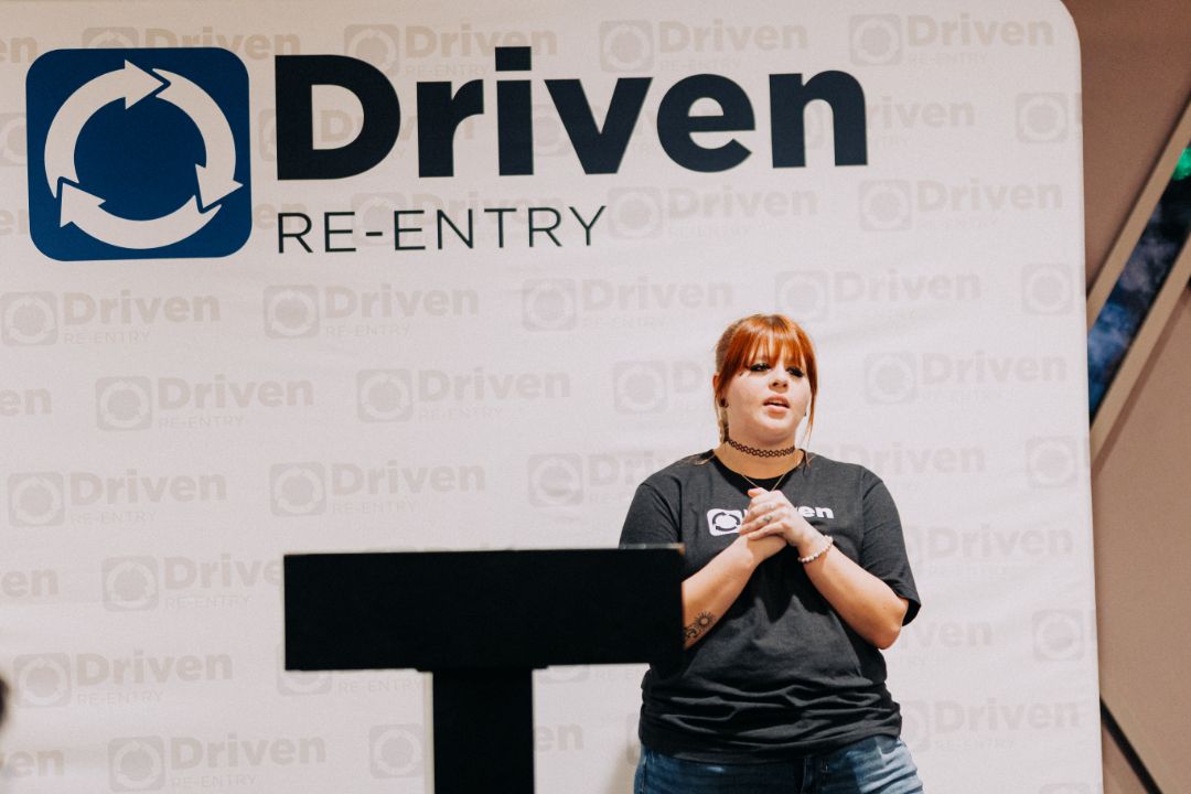 A woman stands in front of a sign that says driven re entry