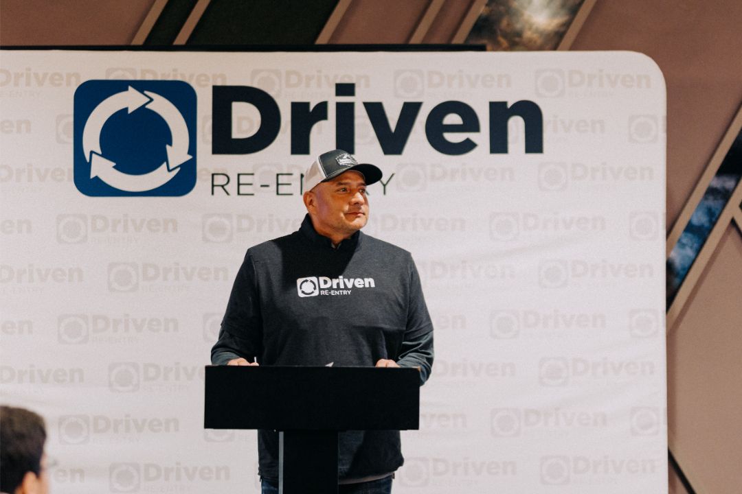 A man stands at a podium in front of a sign that says driven