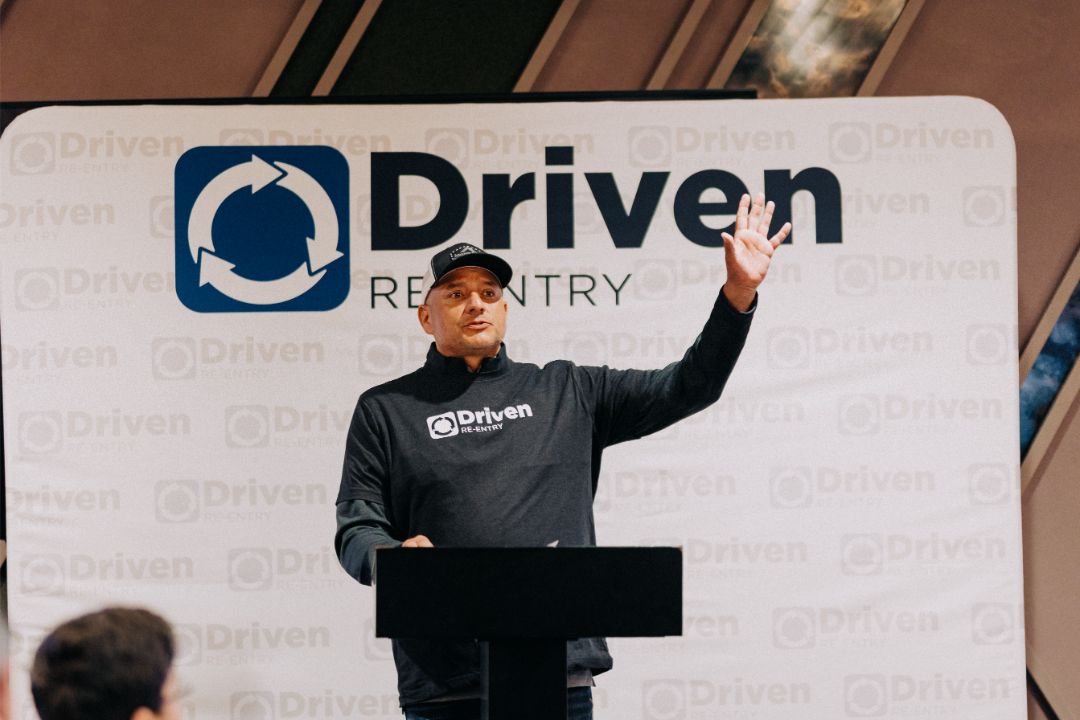 A man is standing at a podium in front of a sign that says driven