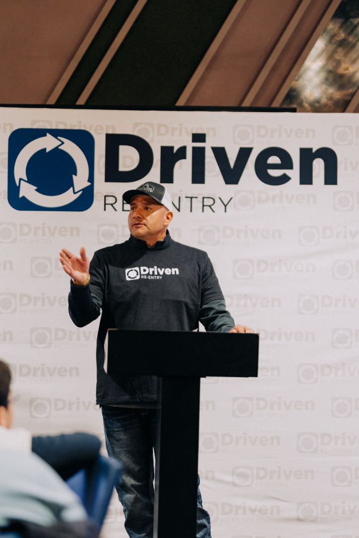 A man stands at a podium in front of a sign that says driven