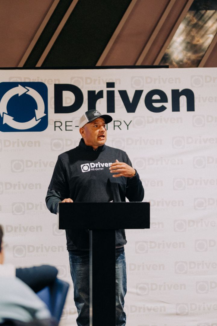 A man stands at a podium in front of a sign that says driven